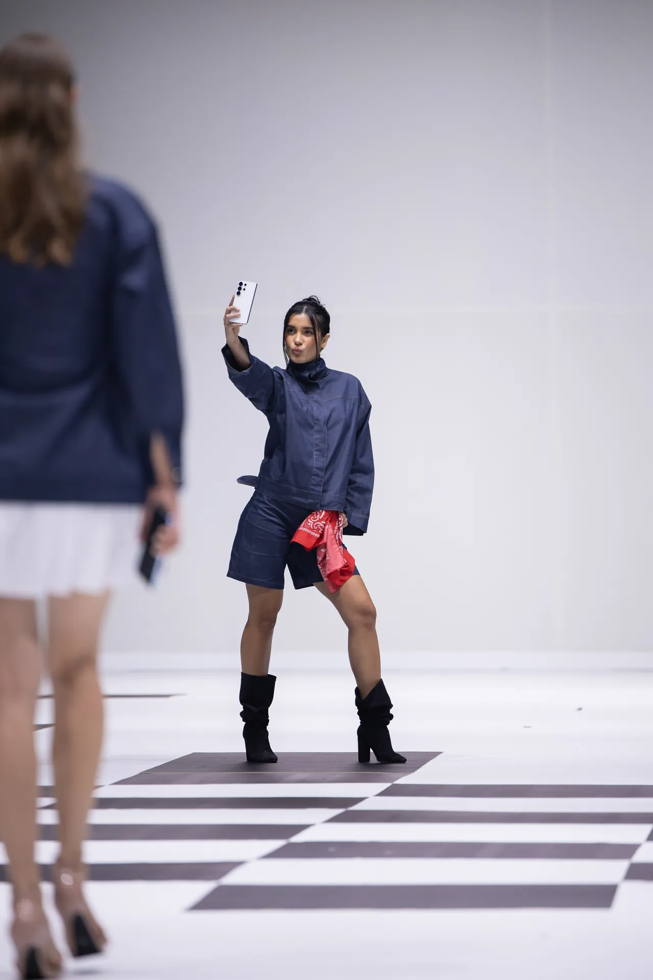 Model on a striped runway taking a selfie with a white smartphone, wearing a dark blue outfit and black boots, red bandana at the waist.