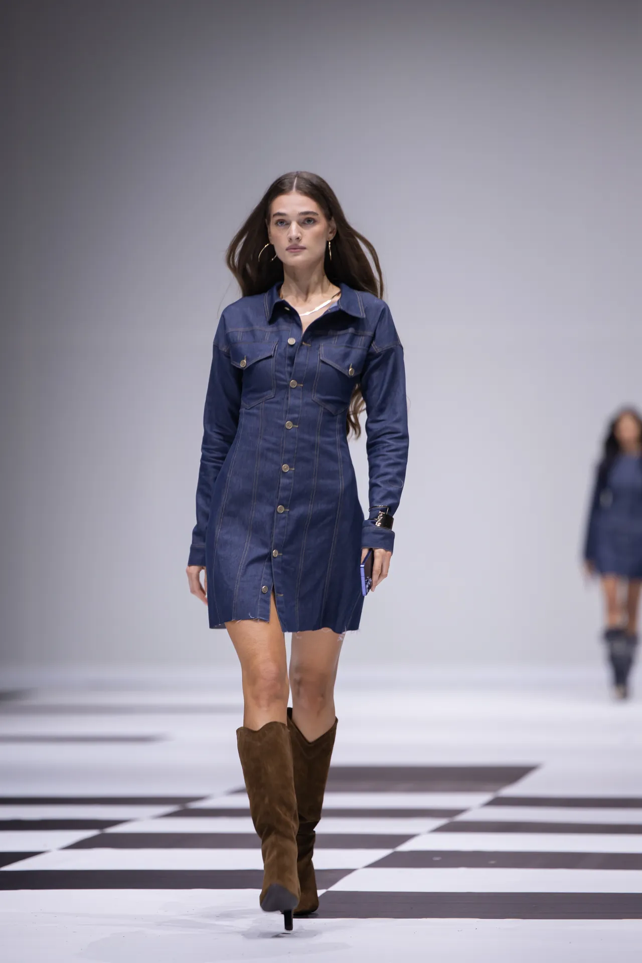 Model walking a fashion runway in a dark denim shirt-dress with brown knee-high boots on a black-and-white checkered floor.