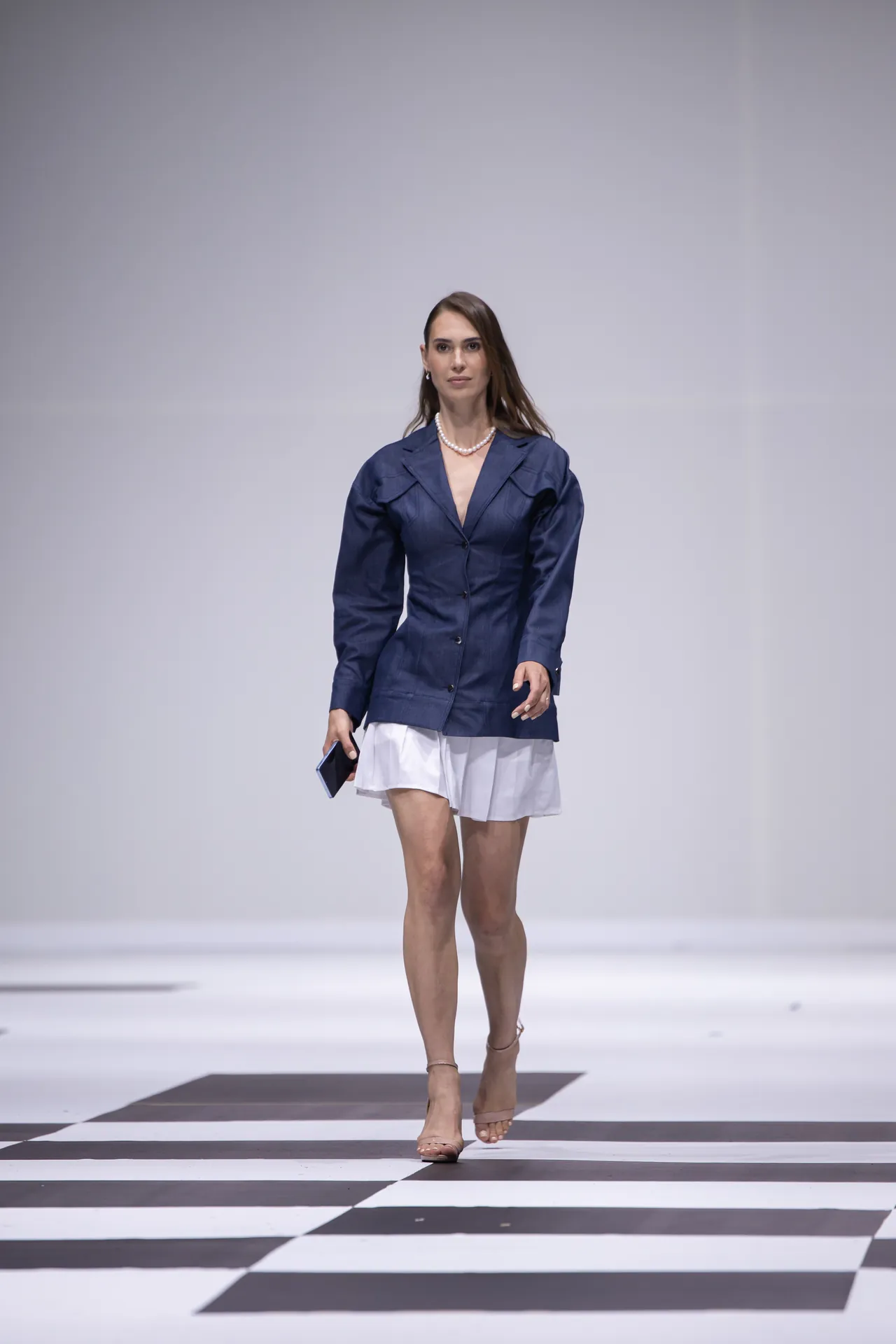 Model walks a fashion runway wearing a navy blazer and white skirt, accessorized with pearls and nude heels, holding a phone.