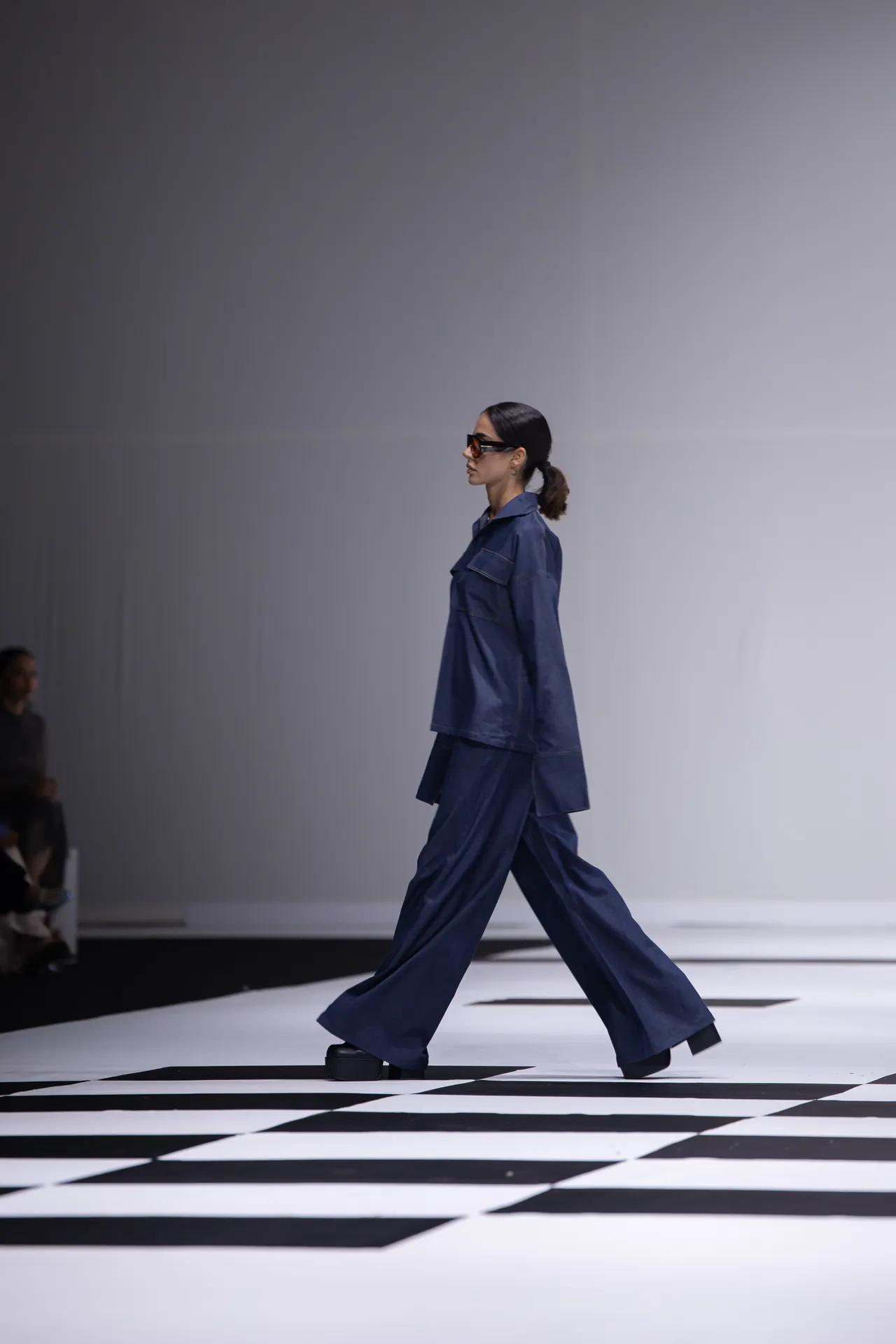 Model walks the runway in a navy blue, oversized pantsuit with sunglasses on a black-and-white checkerboard floor.