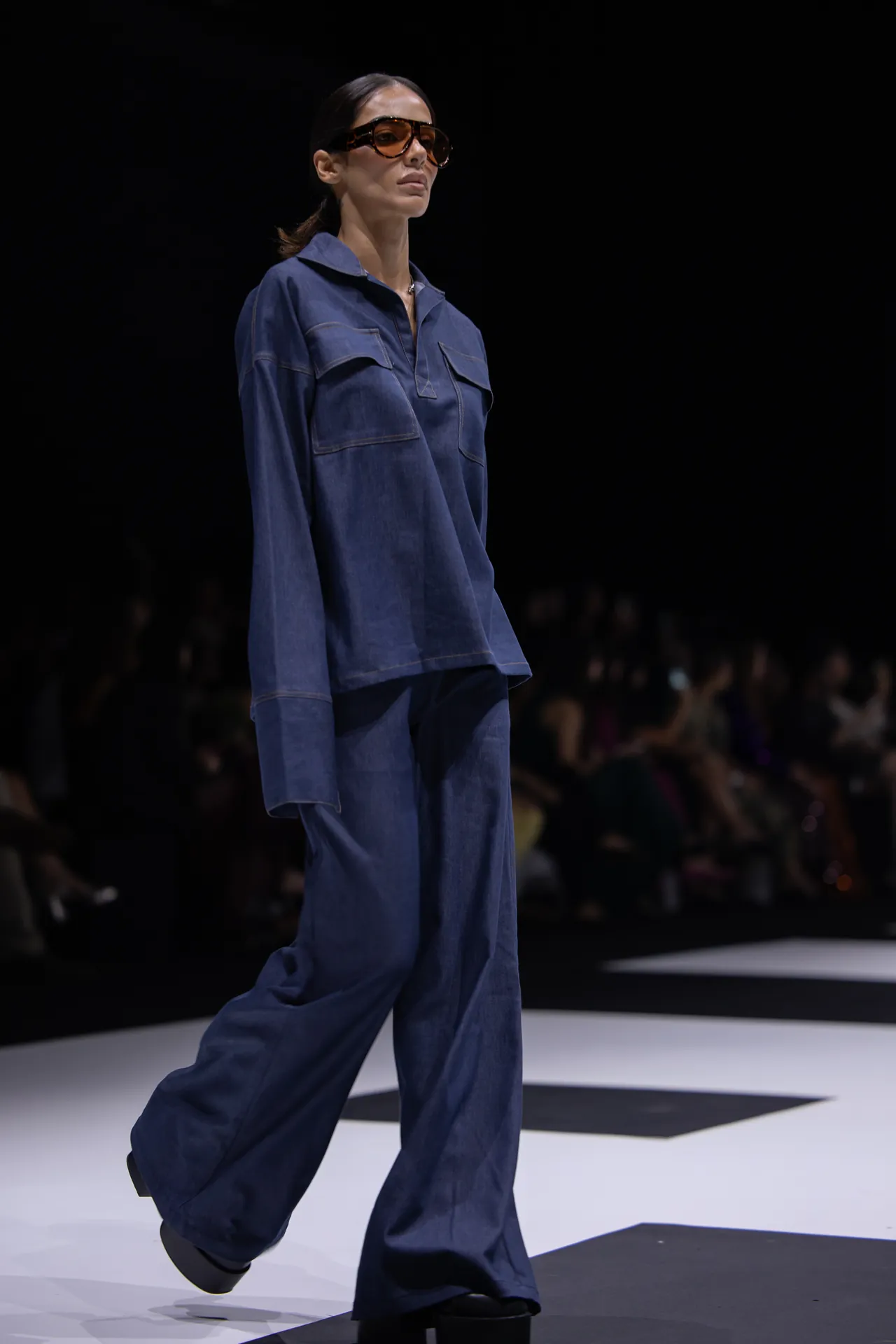 Model walking a fashion runway in a loose blue denim outfit with oversized pockets and brown sunglasses.
