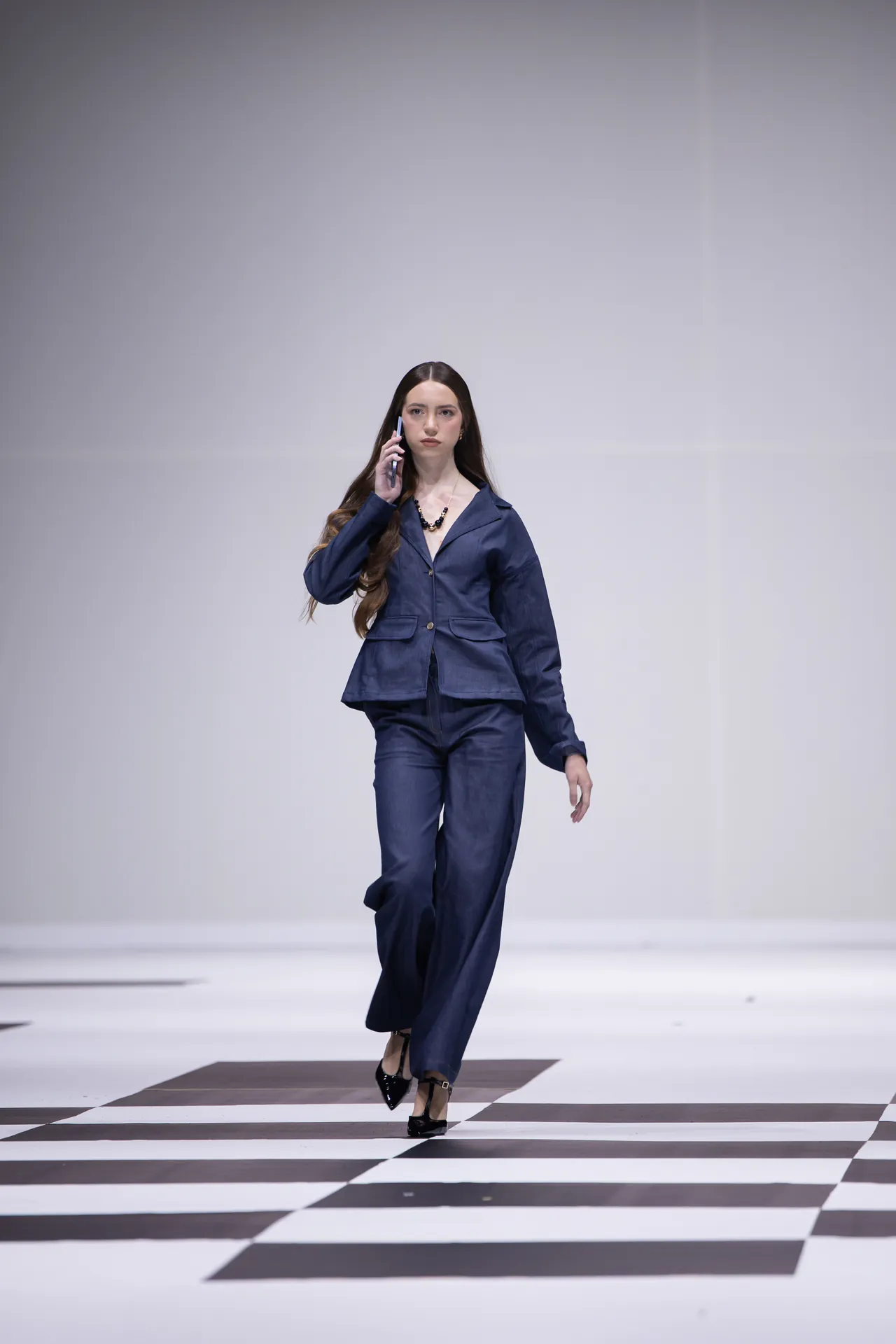 Female model in a navy pantsuit walking down a black-and-white checkered runway, talking on a cellphone at midstride.