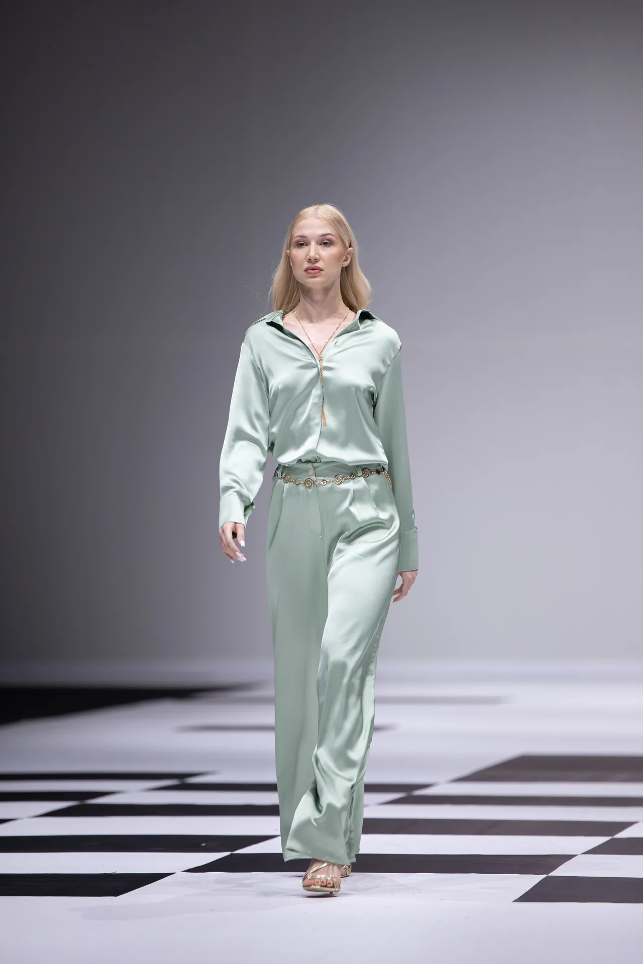 Model walking a fashion runway in a mint-green satin blouse and matching wide-leg pants with a gold belt and nude sandals.