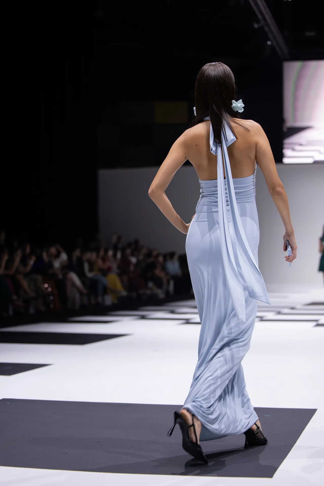 Back view of a model walking the runway in a pale blue halter gown, audience visible in the background.