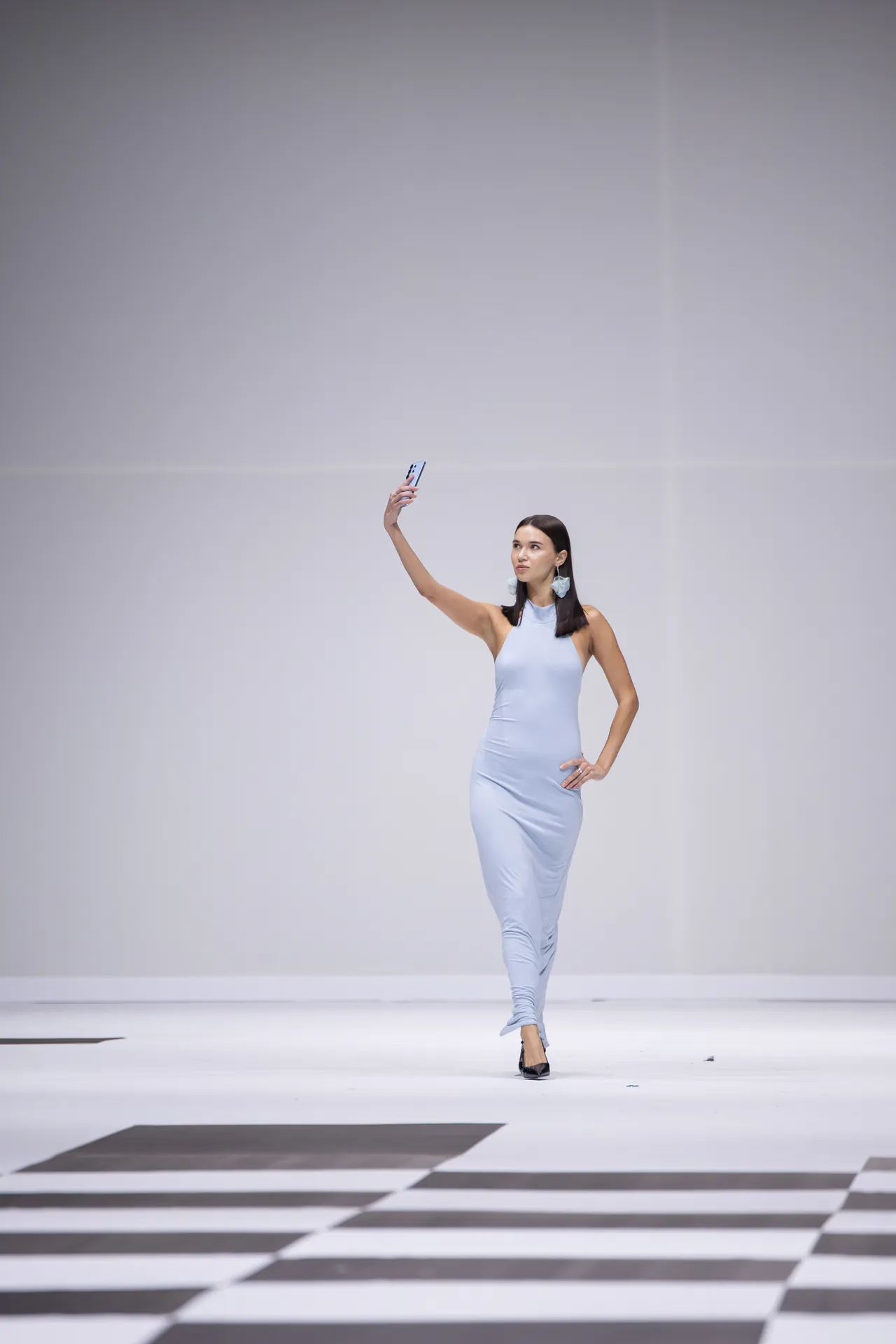 Fashion model in a light blue sleeveless dress posing on a white runway while taking a selfie with her phone.