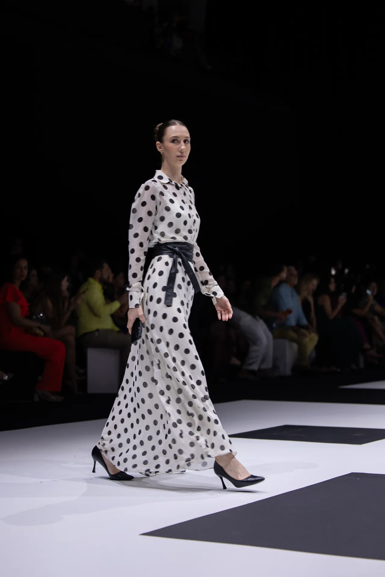 Model walking a fashion runway in a white polka-dot maxi dress with a black sash and black heels, audience in the background.