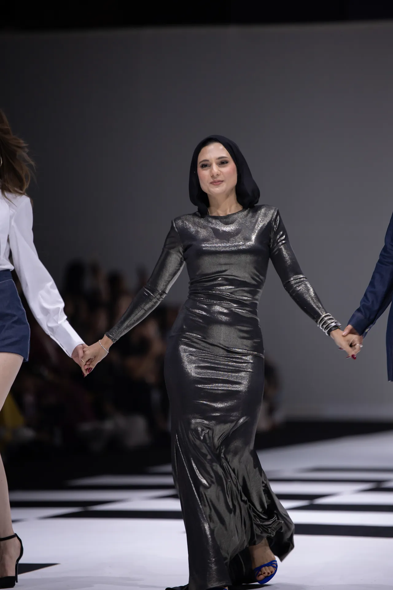 Model in a metallic silver gown and black headscarf walking a fashion show, arms linked with other models on a checkered runway.