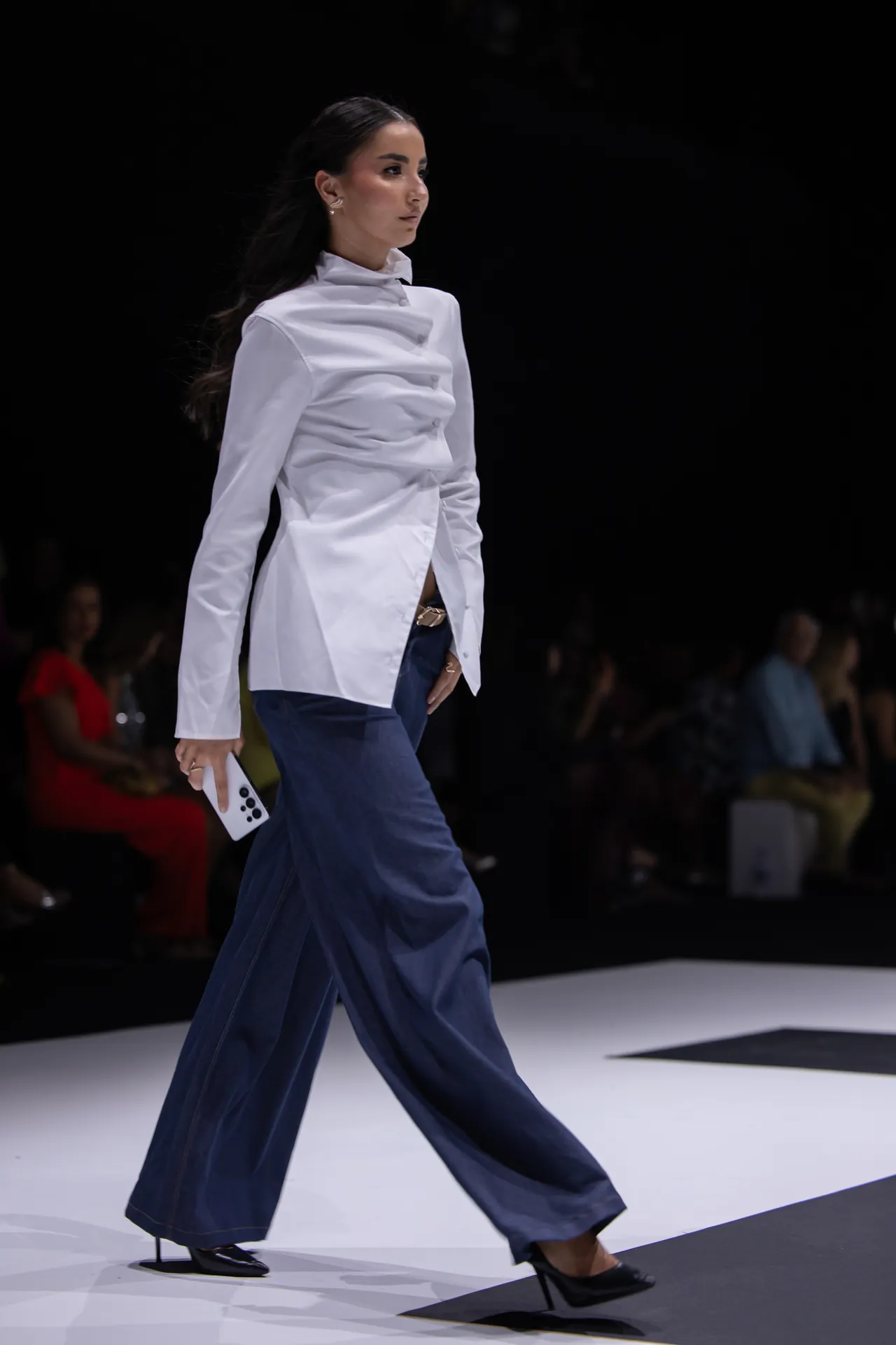 Female model walks the runway in a white structured blouse and navy wide-leg pants, holding a white smartphone at her side.