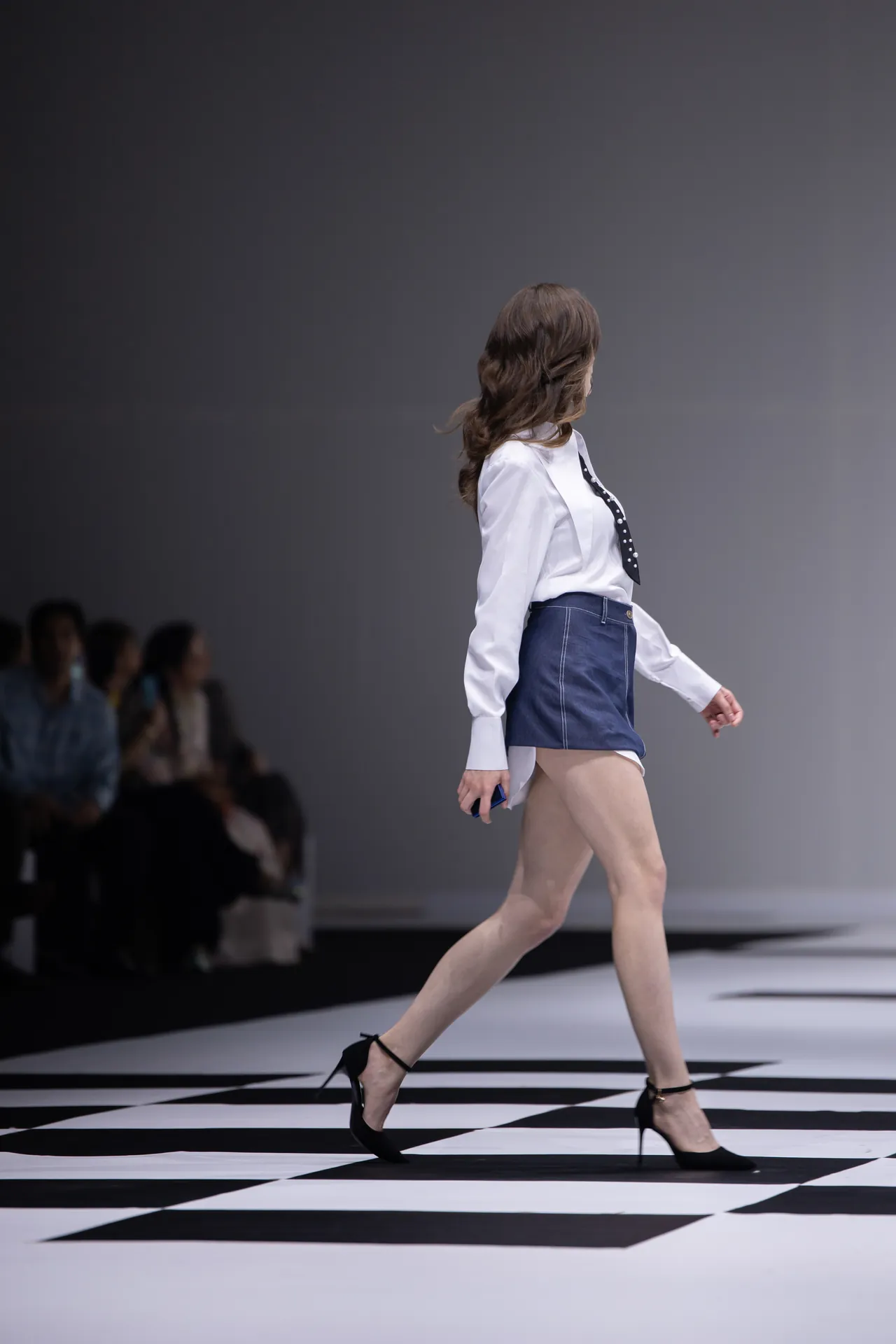 Model walking the runway in a white blouse, dark tie, and blue high-waisted shorts, on a black-and-white checkered floor with an audience in the background.