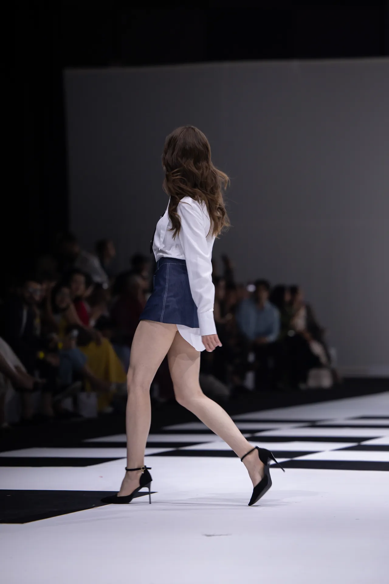 Model walking the runway in a white blouse, blue mini skirt, and black high heels on a black-and-white checkered catwalk.