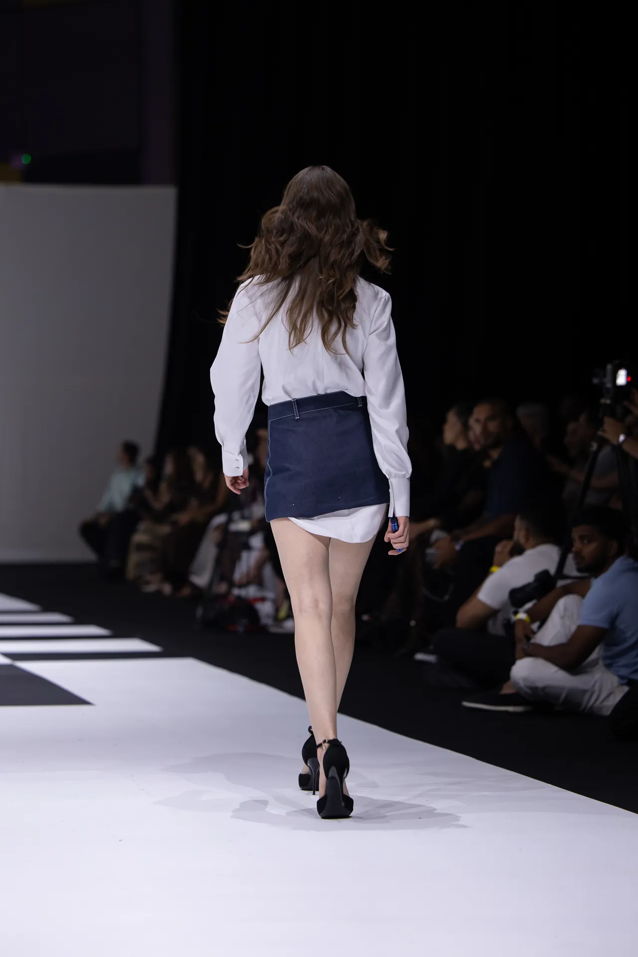 Model walking away on a fashion runway, back view, wearing a white blouse with a dark navy mini skirt over white shorts and black high heels, audience seated along the catwalk