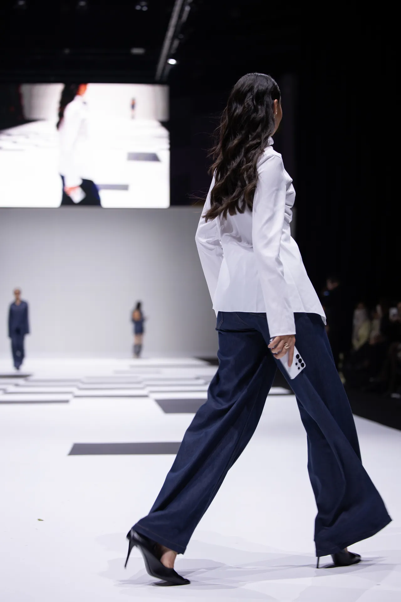 Model walking a fashion runway, back view, wearing a white blouse and navy wide-leg pants, holding a smartphone.