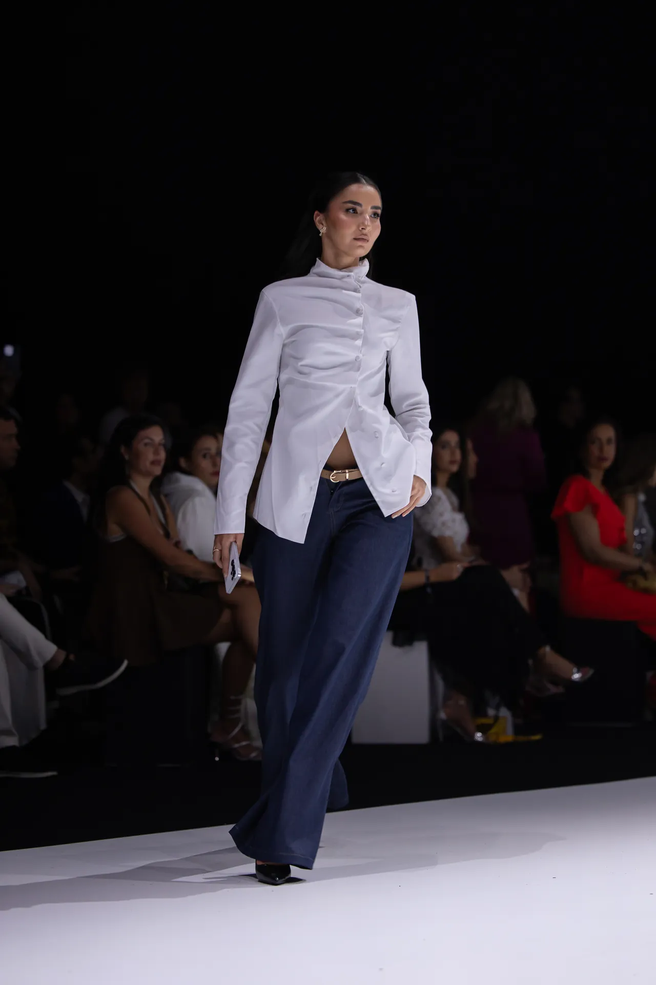 Model walks a fashion runway in a white high-neck blouse with asymmetric button closure and dark blue wide-leg pants, audience in background.