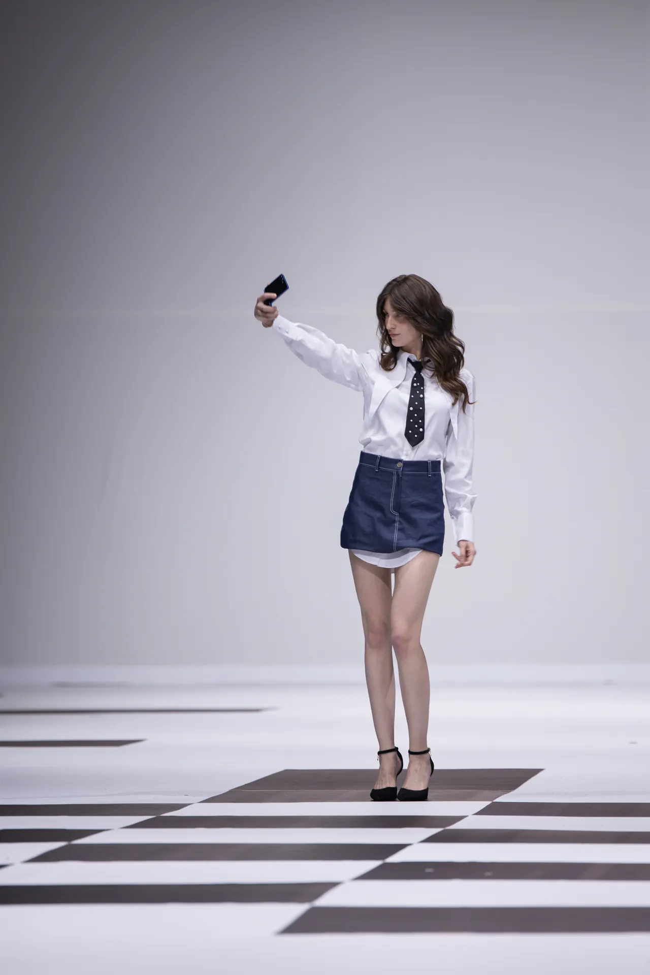 Model on a runway posing and taking a selfie with a smartphone, wearing a white shirt, polka-dot tie, navy mini skirt, and black heels on a black-and-white checkered floor