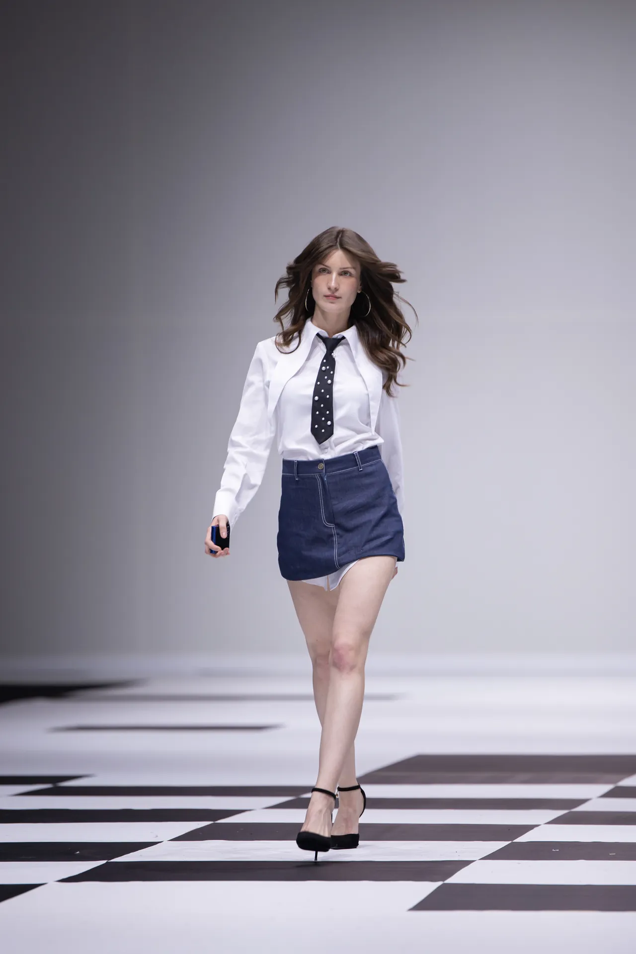 Female модель walking down a runway in a white shirt, dark tie, blue denim miniskirt, black heels, holding a small blue object in her right hand.