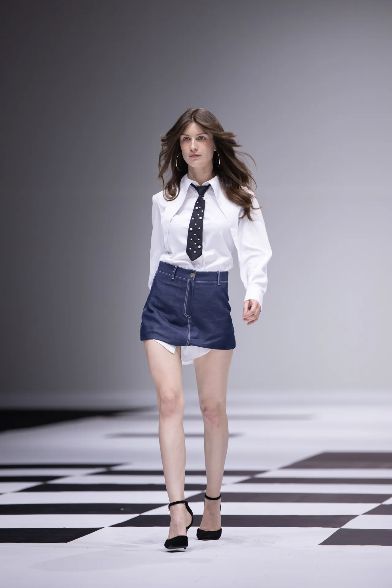 Model walking the runway in a white blouse, polka-dot tie, and navy mini skirt, with black heels on a black-and-white checkered floor.