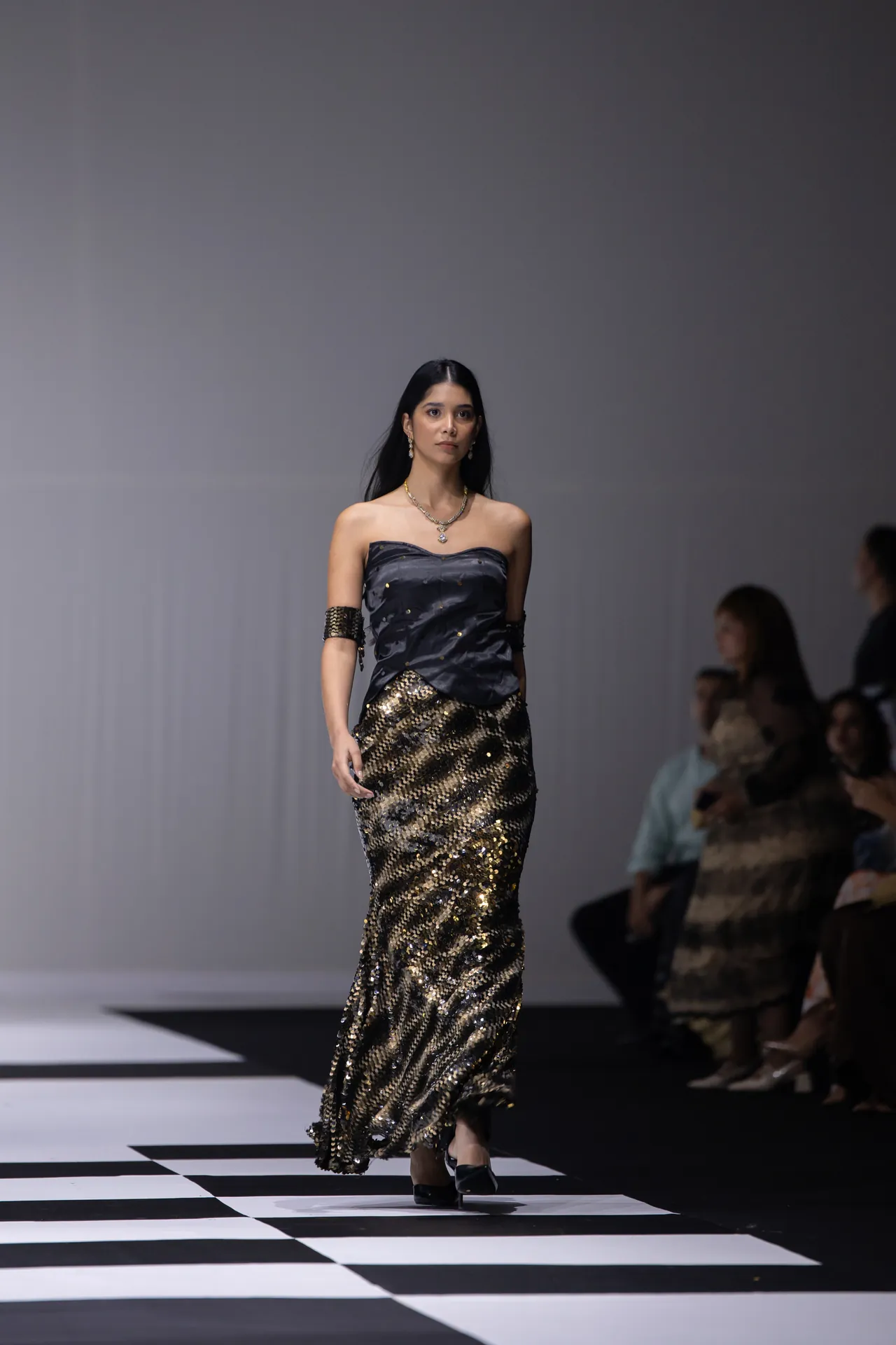 Female model walking a black-and-gold sequined strapless gown on a black-and-white checkered runway, accessorized with jewelry and black heels.