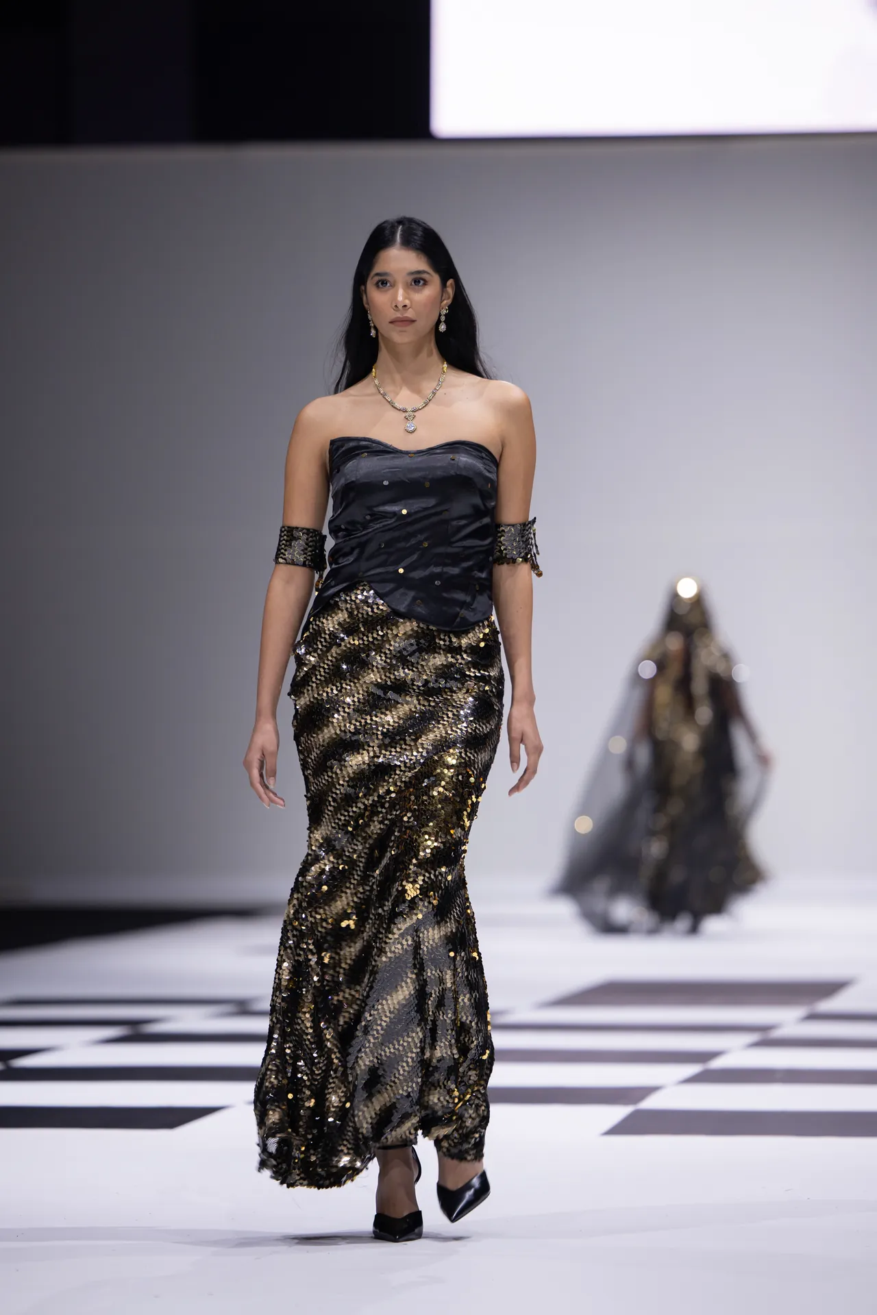Model walking the runway in a strapless black bodice and a gold sequined mermaid-style skirt, accessorized with jewelry.