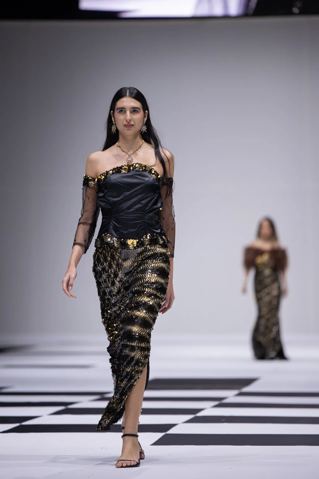 Model walking a runway in a black off-shoulder dress with gold sequins and sheer sleeves, paired with black sandals on a black-and-white checkered floor.