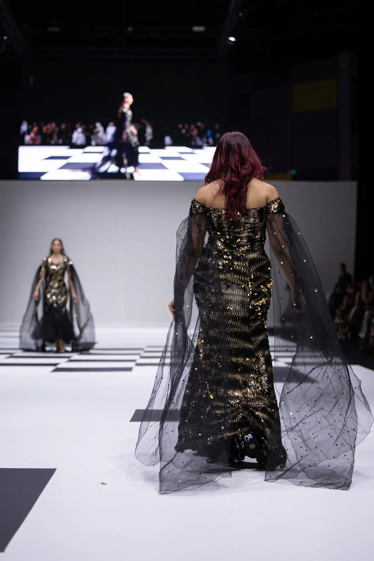 Model in a gold sequin gown with a sheer, dotted cape walks the runway, seen from behind on a black-and-white checker floor.