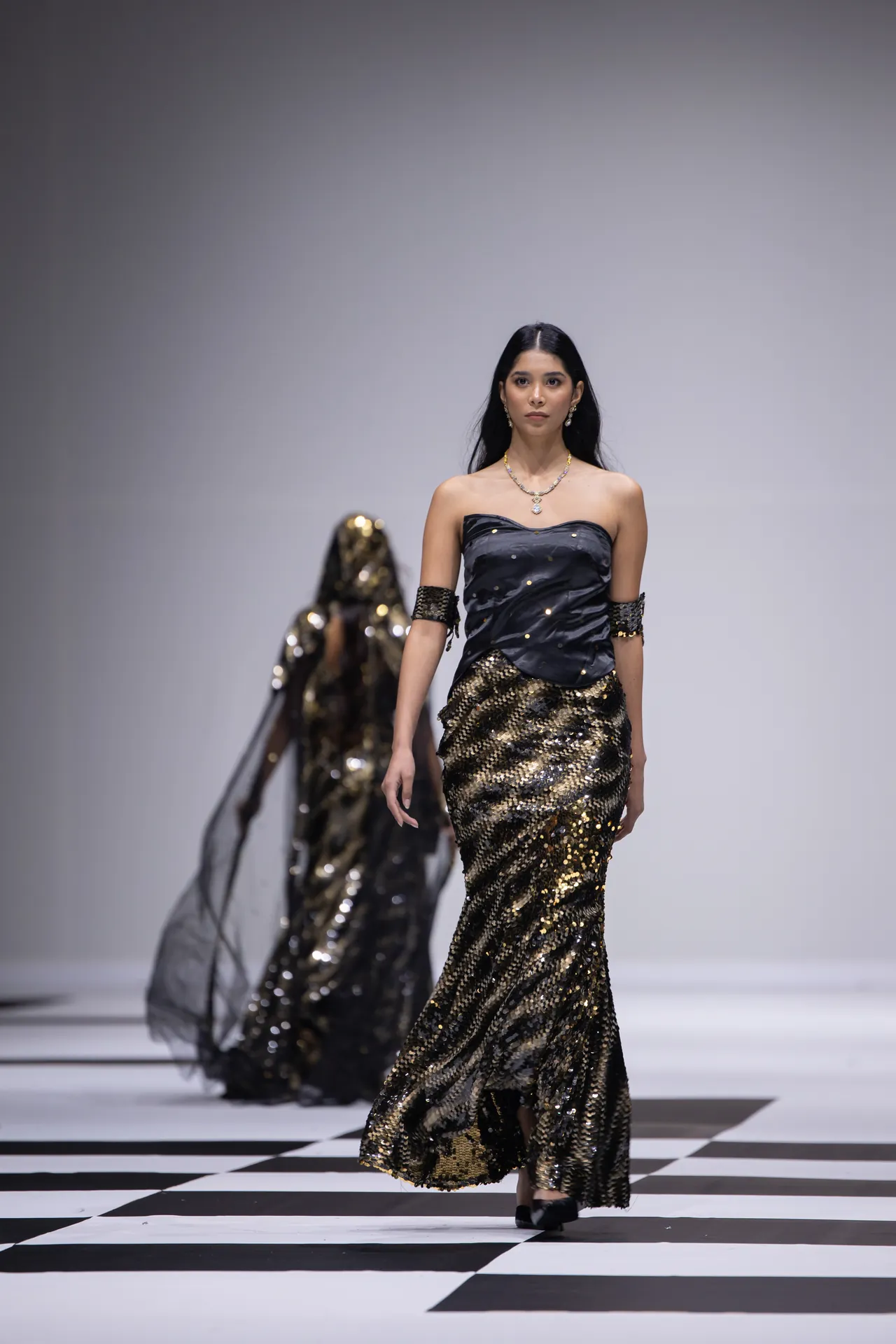 Model walks the runway in a strapless black top and gold sequin mermaid skirt, wearing jewelry on a black-and-white checkered catwalk.