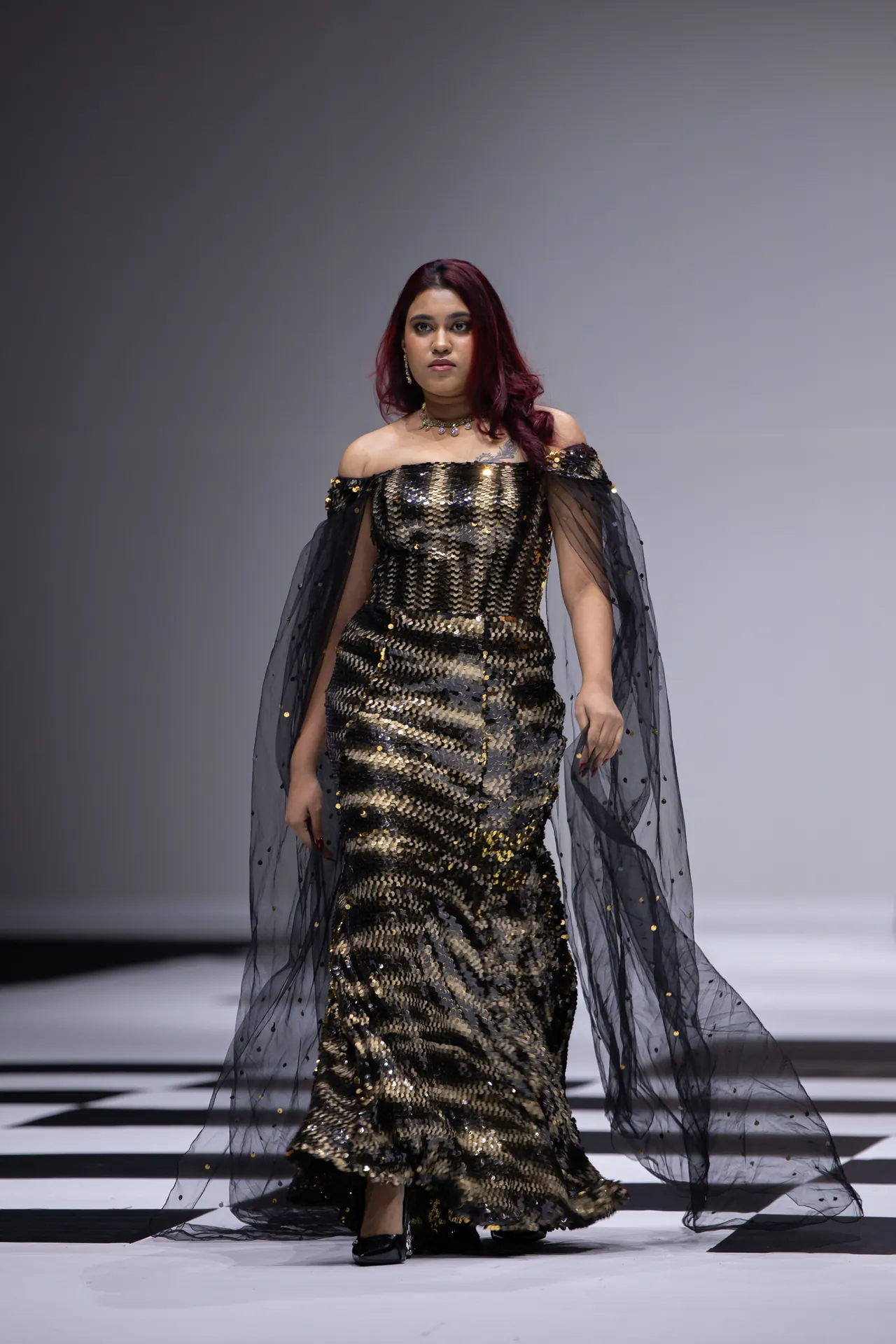 Model walks a runway in a gold sequined off-the-shoulder gown with a sheer black cape adorned with tiny dots.