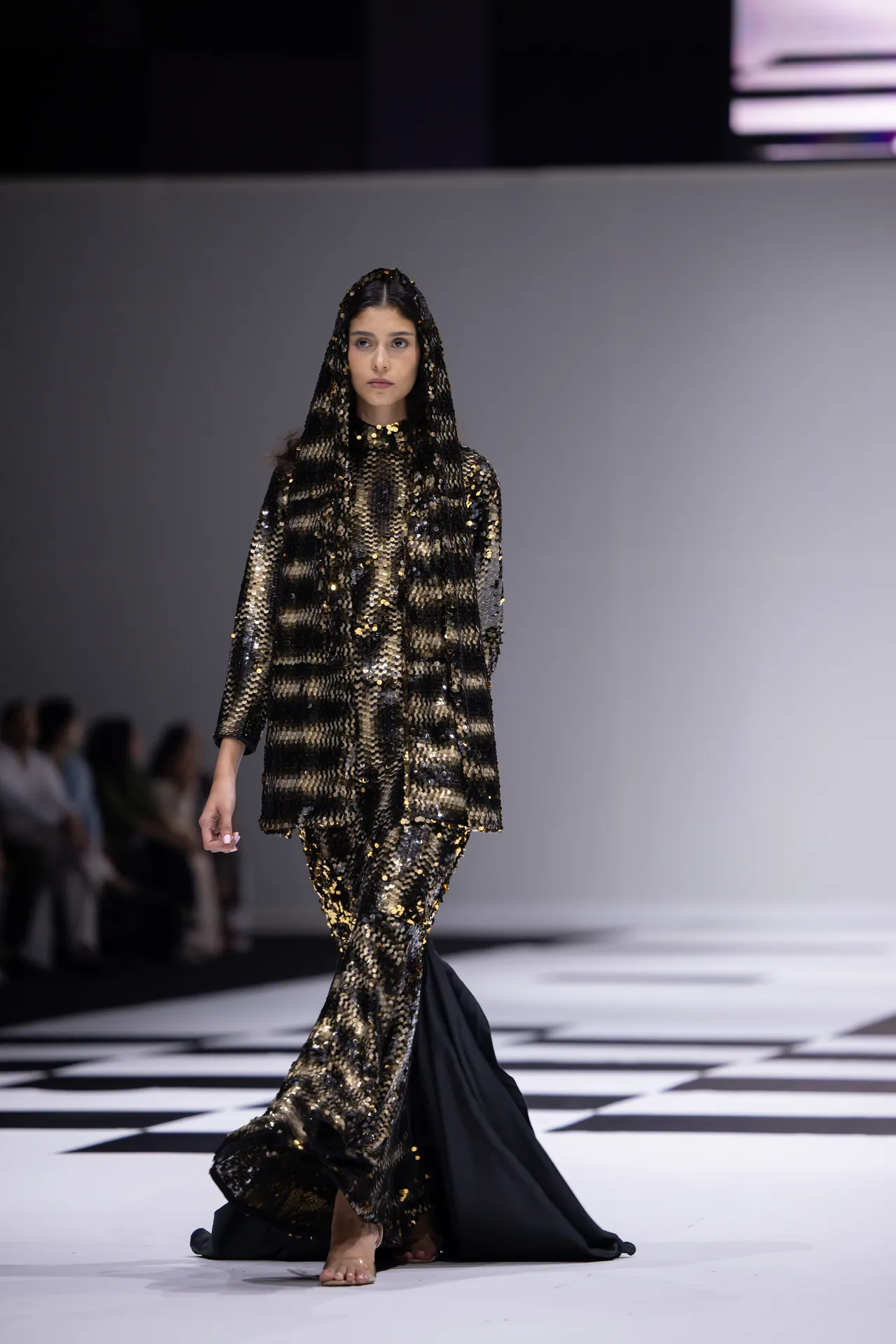 Female model walking a fashion runway in a black-and-gold sequined gown with a long train and hooded cape.