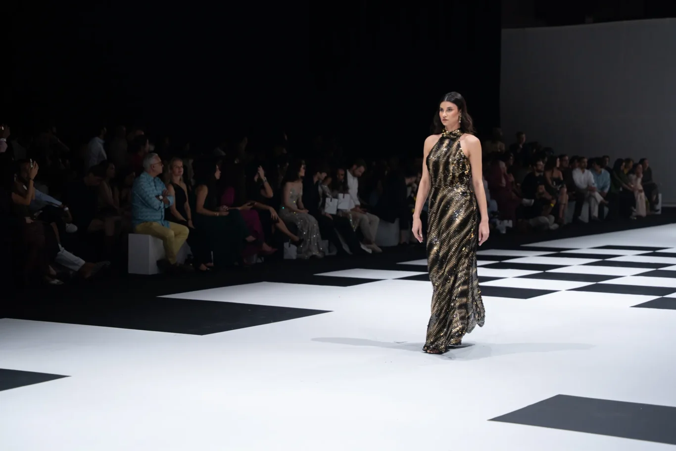 Model walking a gold and black striped gown down a white checkerboard runway with seated spectators at the sides of the catwalk.