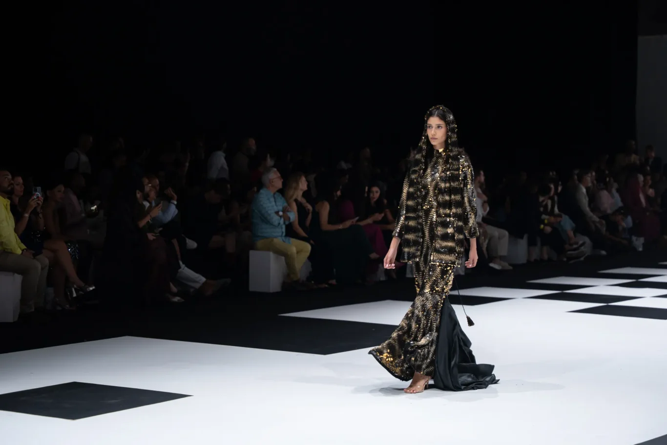 Model strides down a black-and-white checkered runway in a gold sequined hooded coat; seated audience watches in dim lighting.