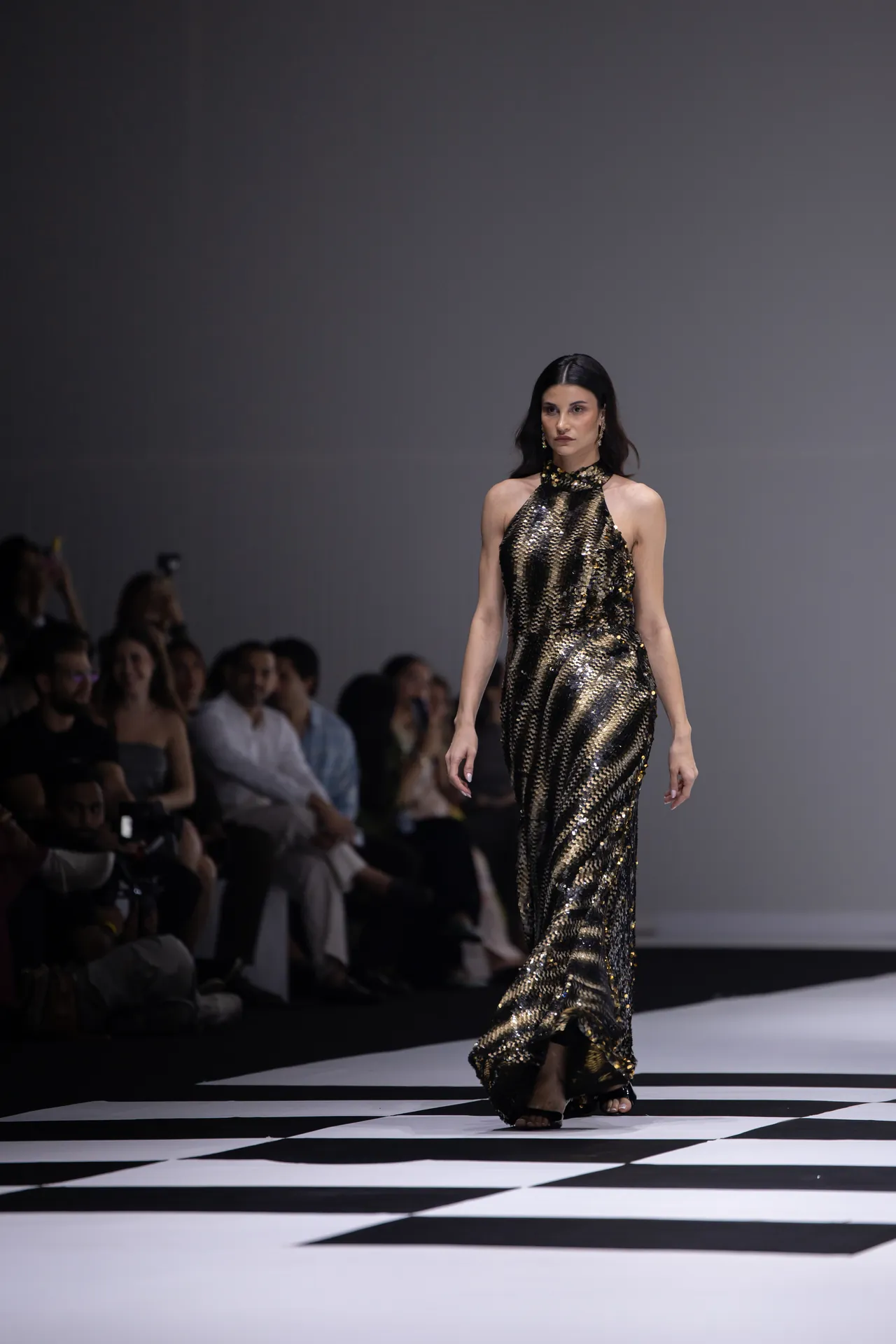 Model wearing a gold and black sequined halter gown walking a fashion runway with the audience in the background on the left.