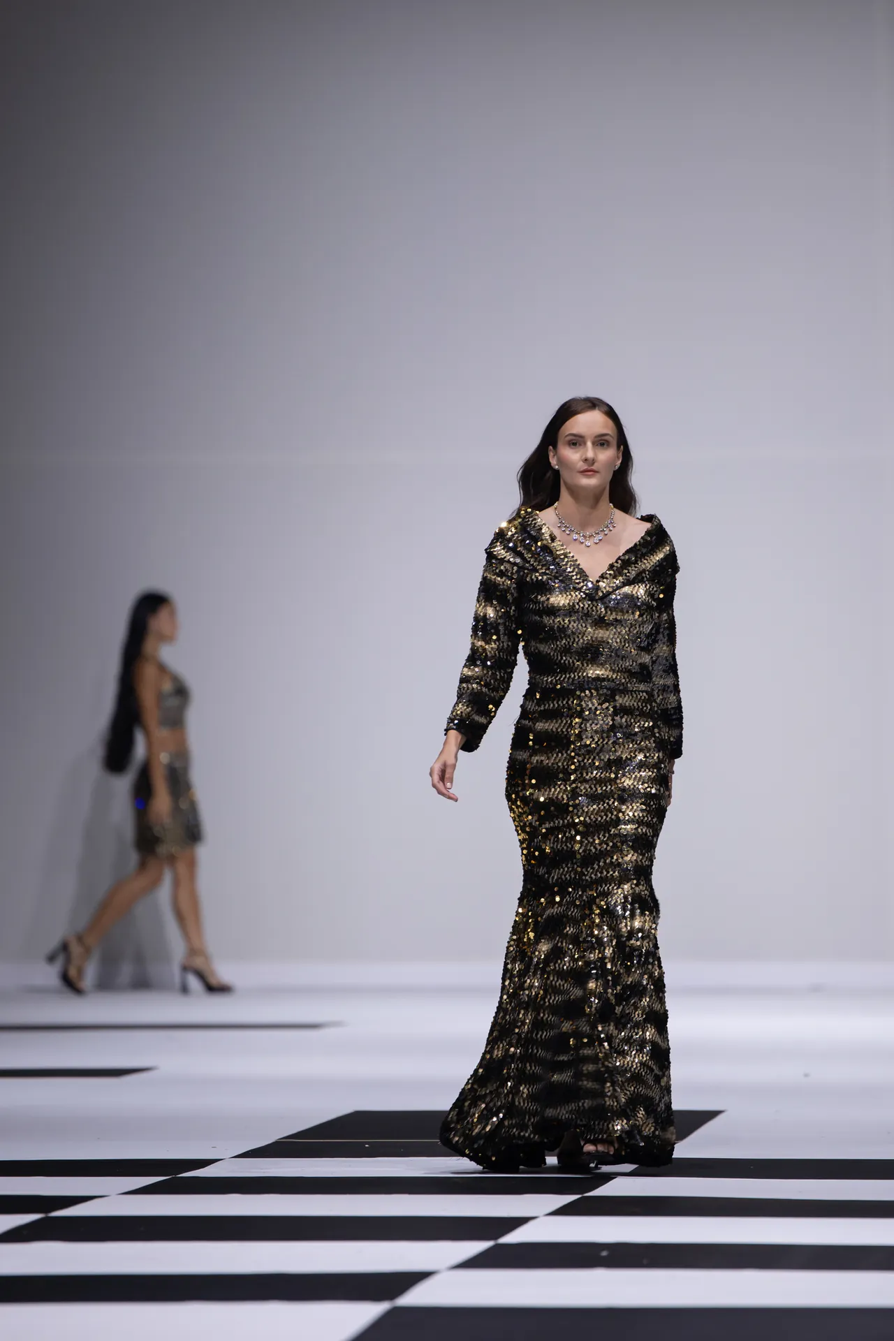 Model walking the runway in a long black-and-gold sequin gown with a necklace on a black-and-white checkered catwalk strengthened by strong lighting.