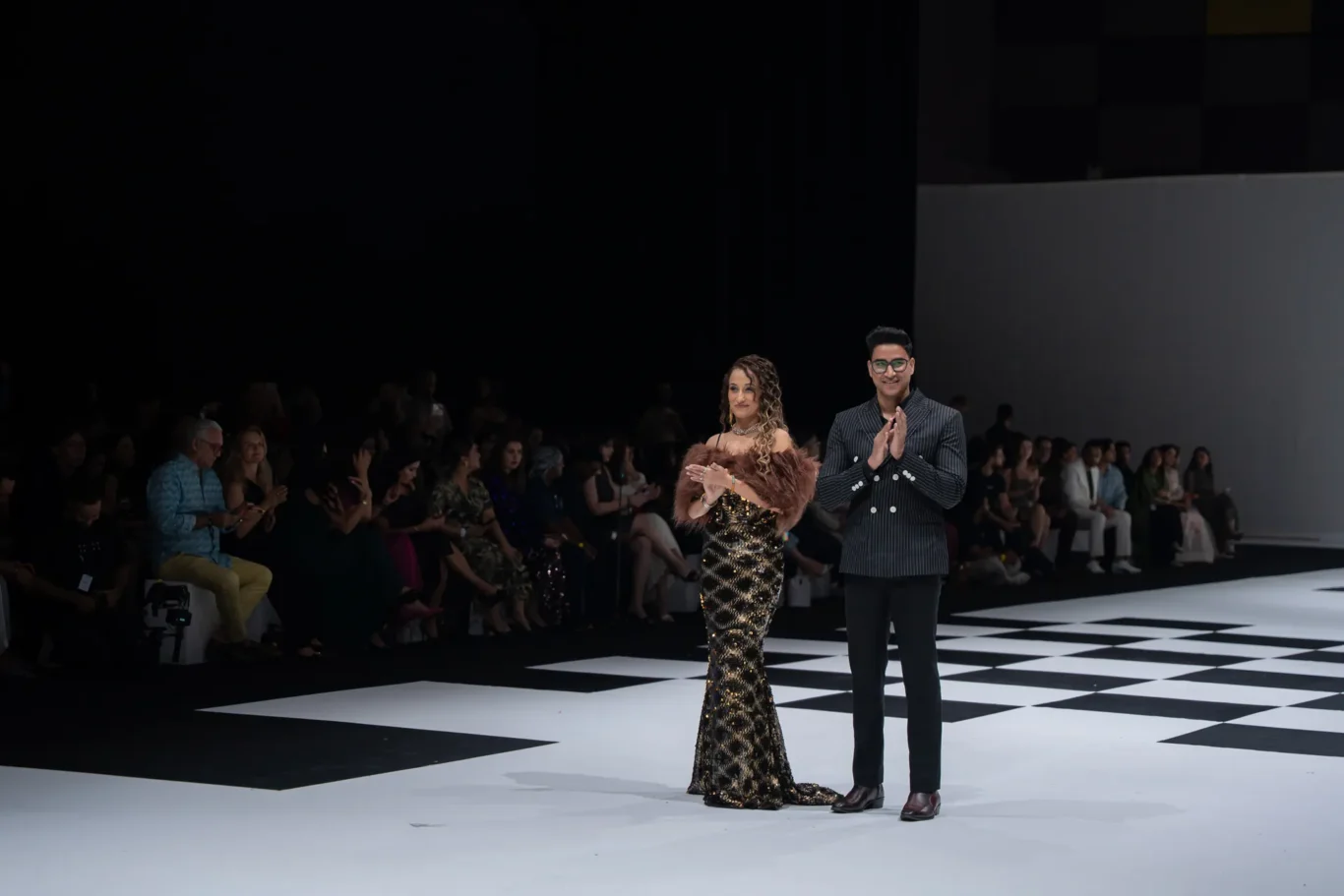 A fashion show runway with a man in a pinstripe suit and a woman in a sequined gown applauding as the models bow. Audience sits along the catwalk in dim lighting.