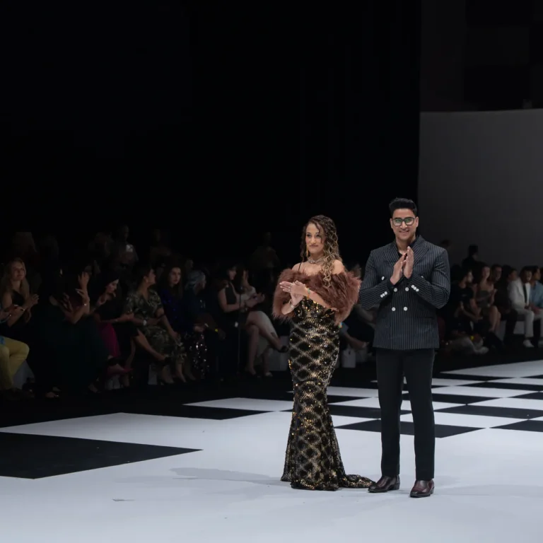 A fashion show runway with a man in a pinstripe suit and a woman in a sequined gown applauding as the models bow. Audience sits along the catwalk in dim lighting.