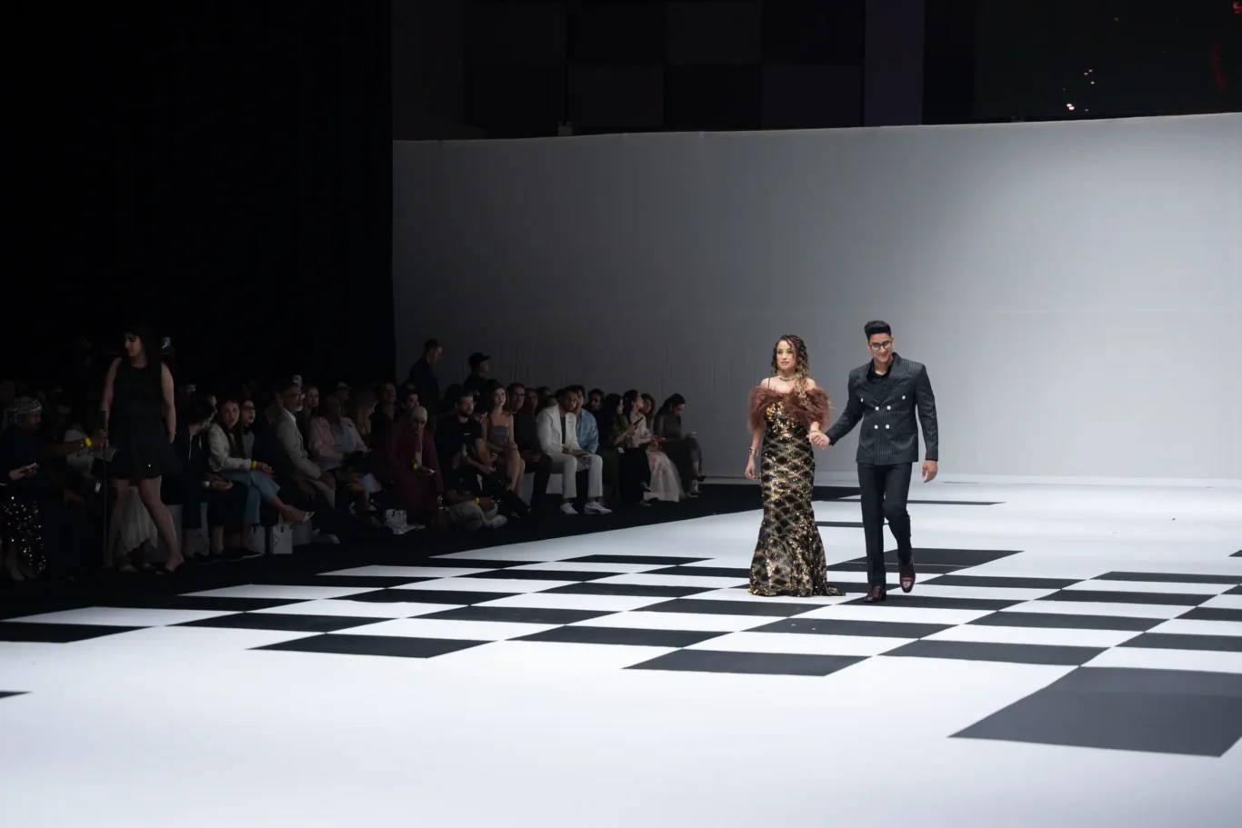 A man and woman in formal evening wear walk hand in hand down a black-and-white checkered runway as a seated audience watches from the sides on a fashion show stage.