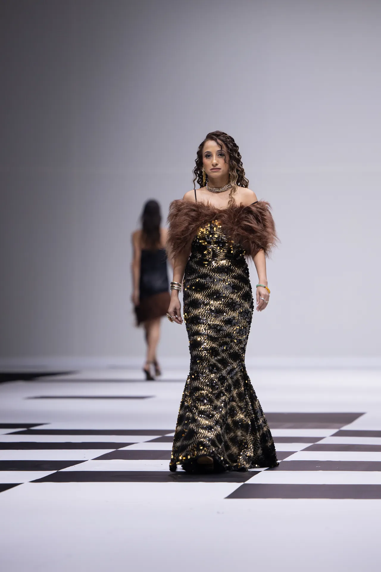 Model wearing a floor-length gold sequined gown with a brown fur stole on a black-and-white checkerboard runway.