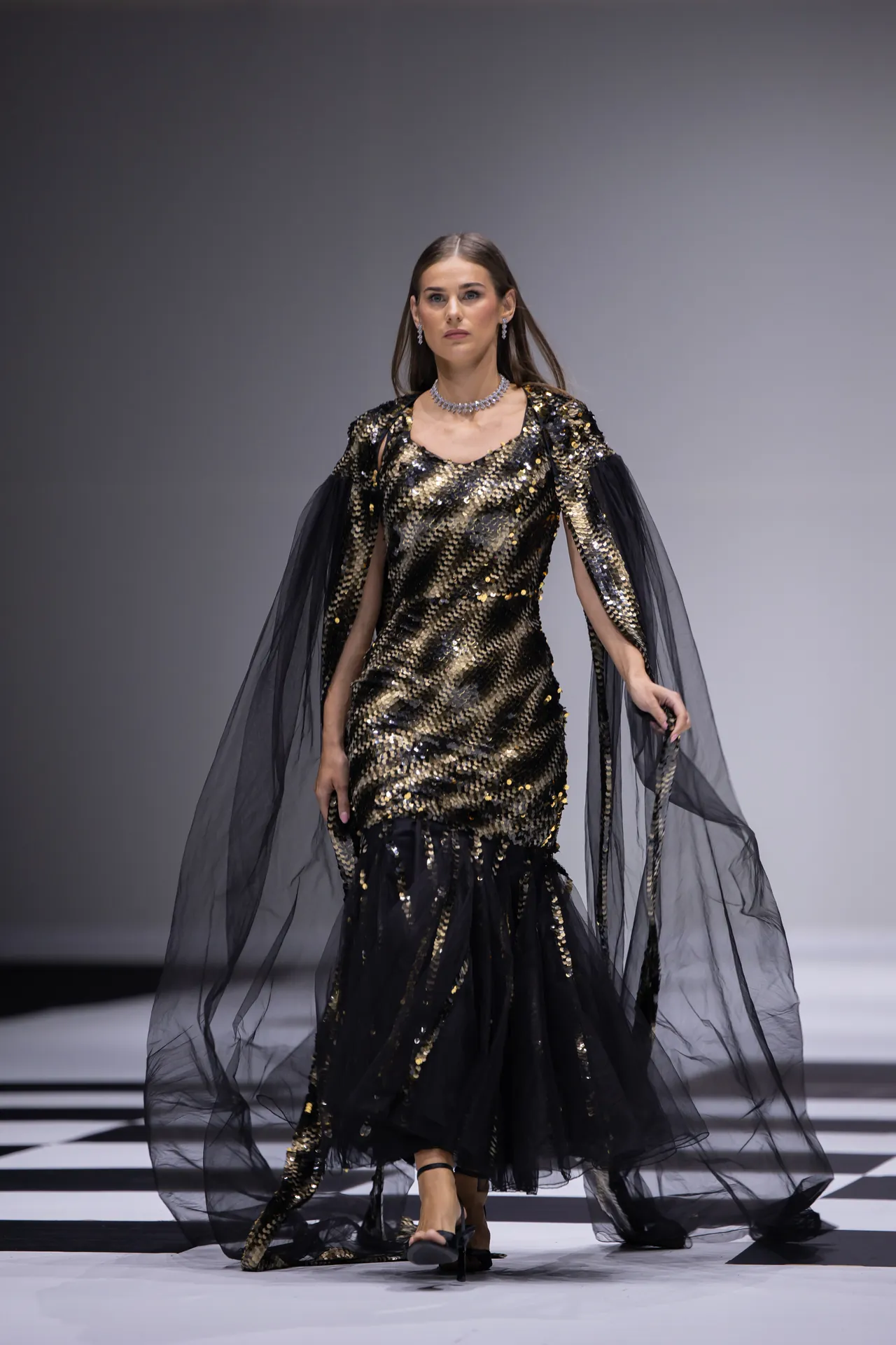 Model walking the runway in a black-and-gold sequin gown with dramatic sheer cape, mid-stride on a checkered runway.