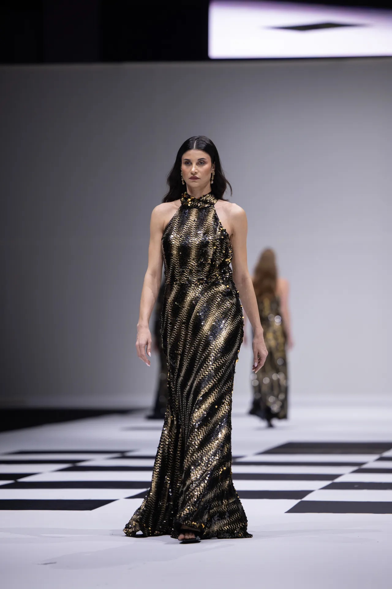 Model wearing a gold and black sequin halter gown walks down a runway with a checkered floor.