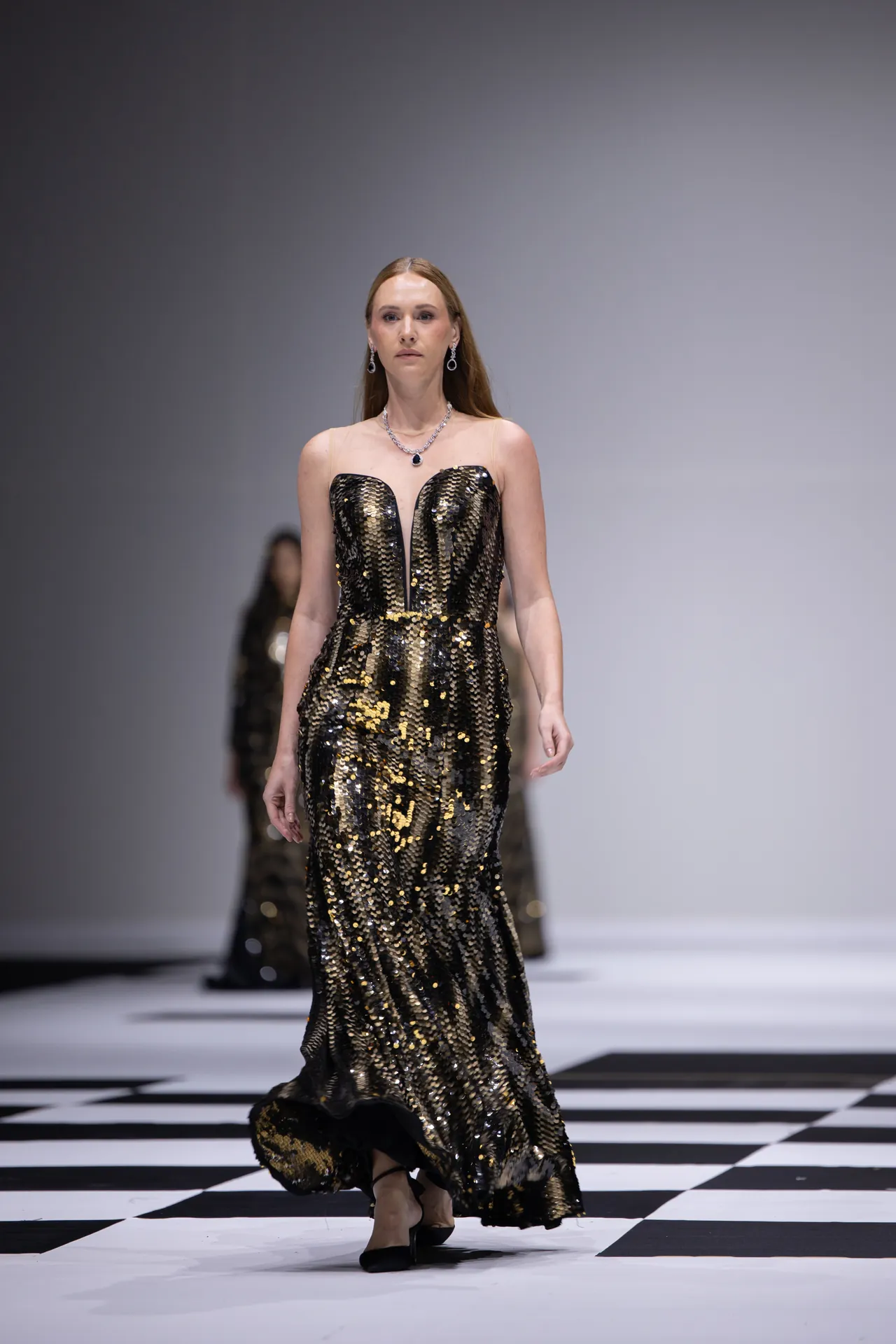 Model walks a runway in a black and gold sequin evening gown, on a checkerboard-patterned floor with a gray backdrop behind her.
