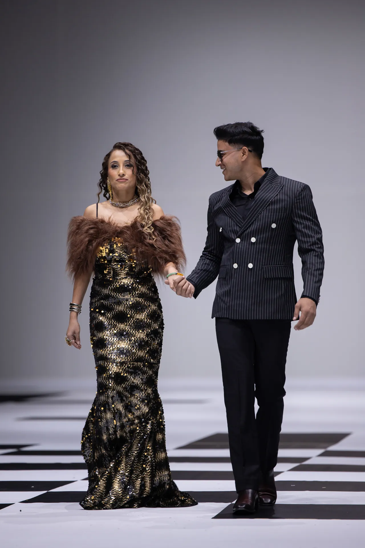 Couple walking hand in hand on a fashion runway; woman wears a gold sequin gown with a fur stole and jewelry, man in a pinstripe suit.
