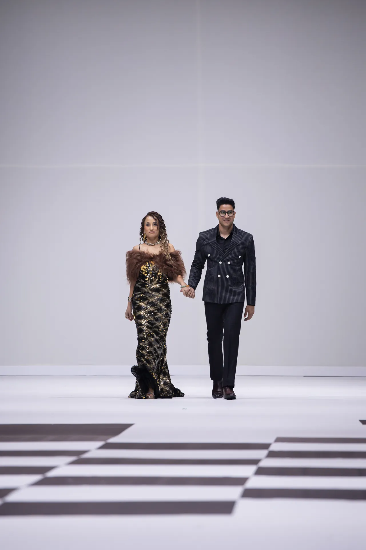 Two fashion models walk hand in hand down a runway, the woman wearing a gold sequin gown with a fur stole and the man in a dark pinstripe suit.