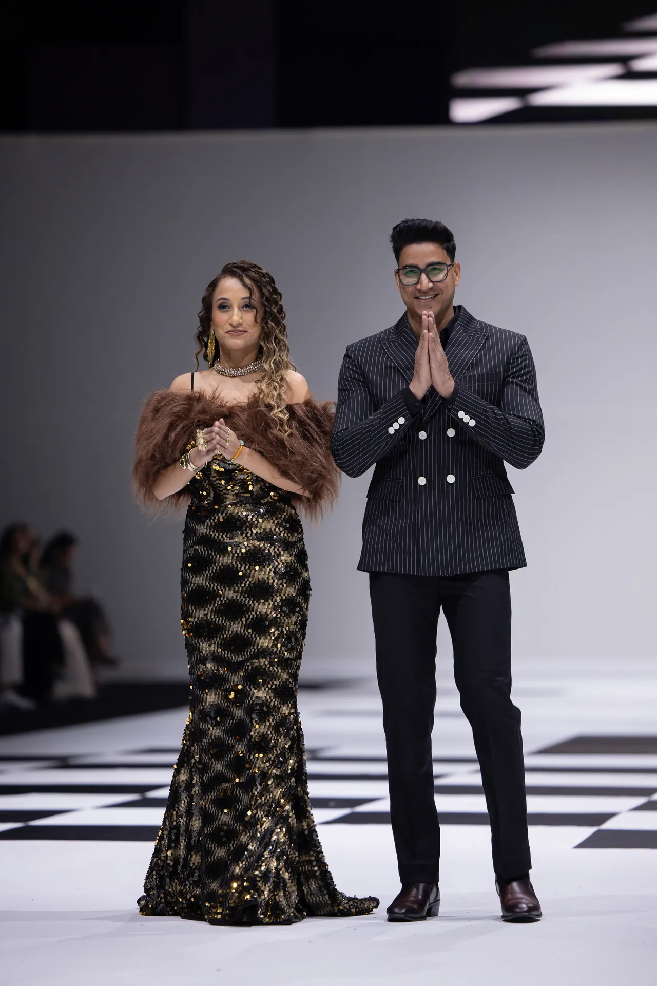 Two models stand on a fashion runway; woman in a gold sequin gown with a fur stole, man in a pinstripe blazer with hands pressed together in front.