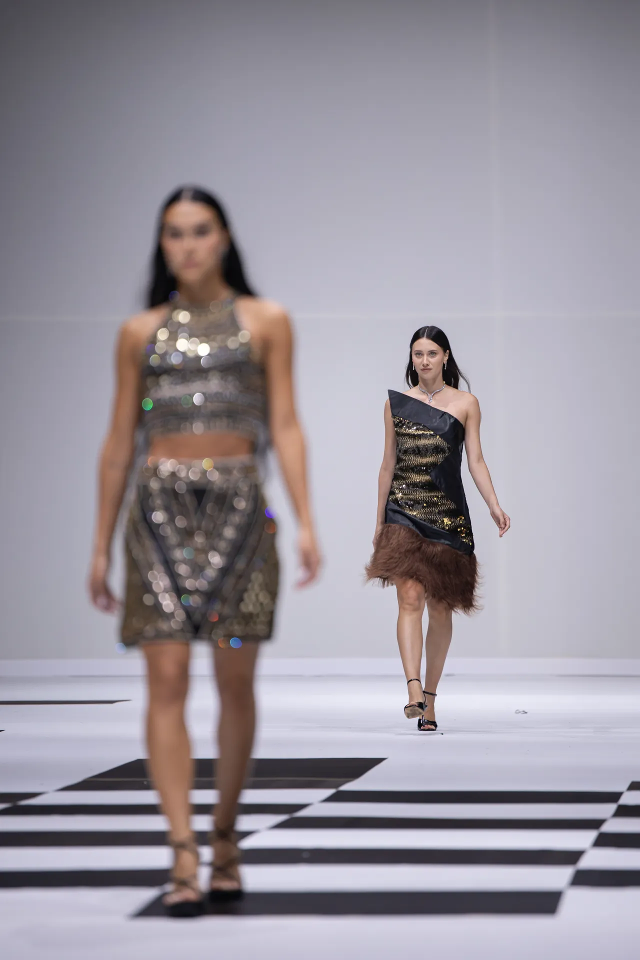 Two models walk a fashion runway in metallic outfits; the foreground model wears a black-and-gold dress, the background model in a shimmering silver ensemble.