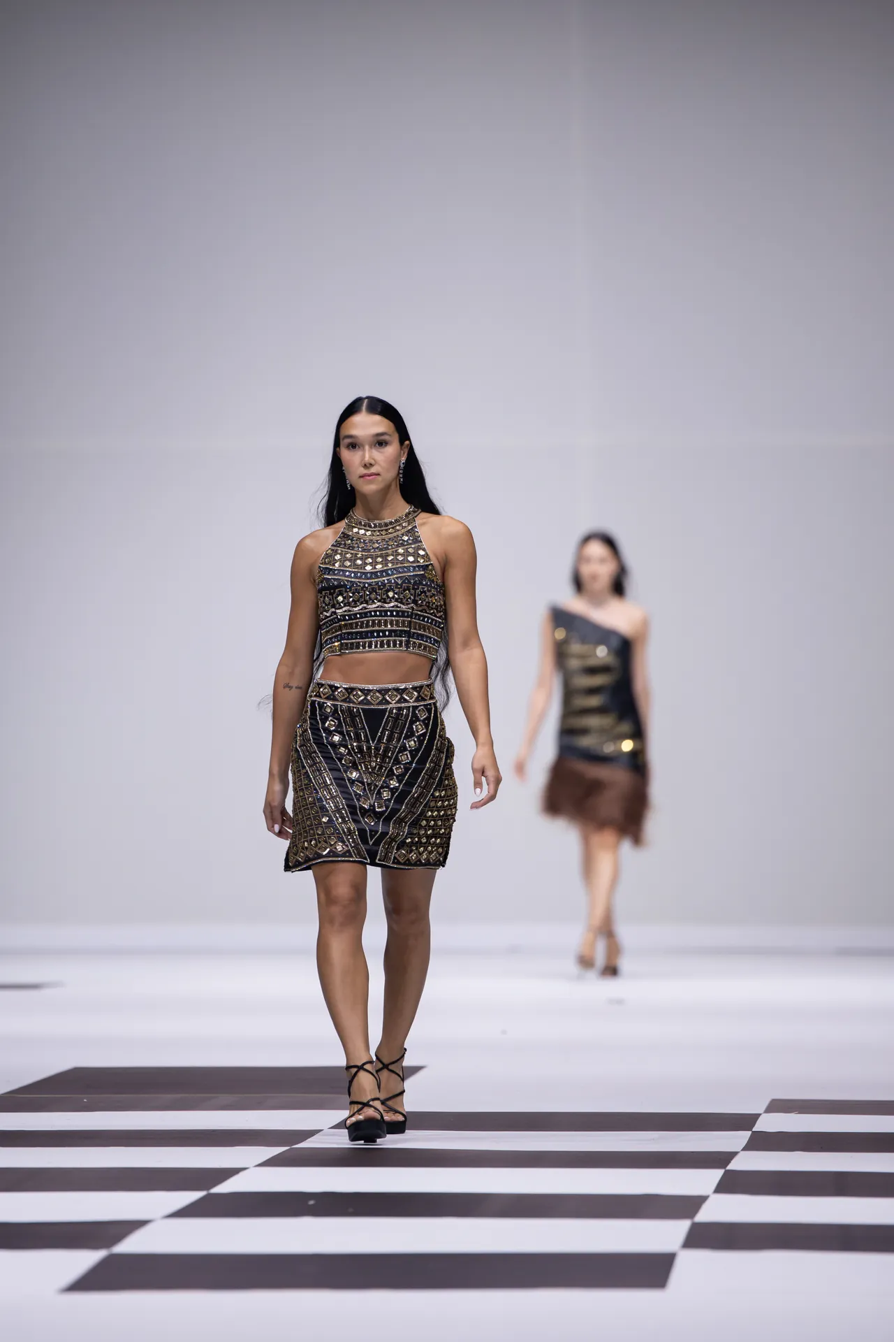 Model walks a fashion runway in a black-and-gold geometric two-piece outfit with strappy heels on a checkerboard catwalk.