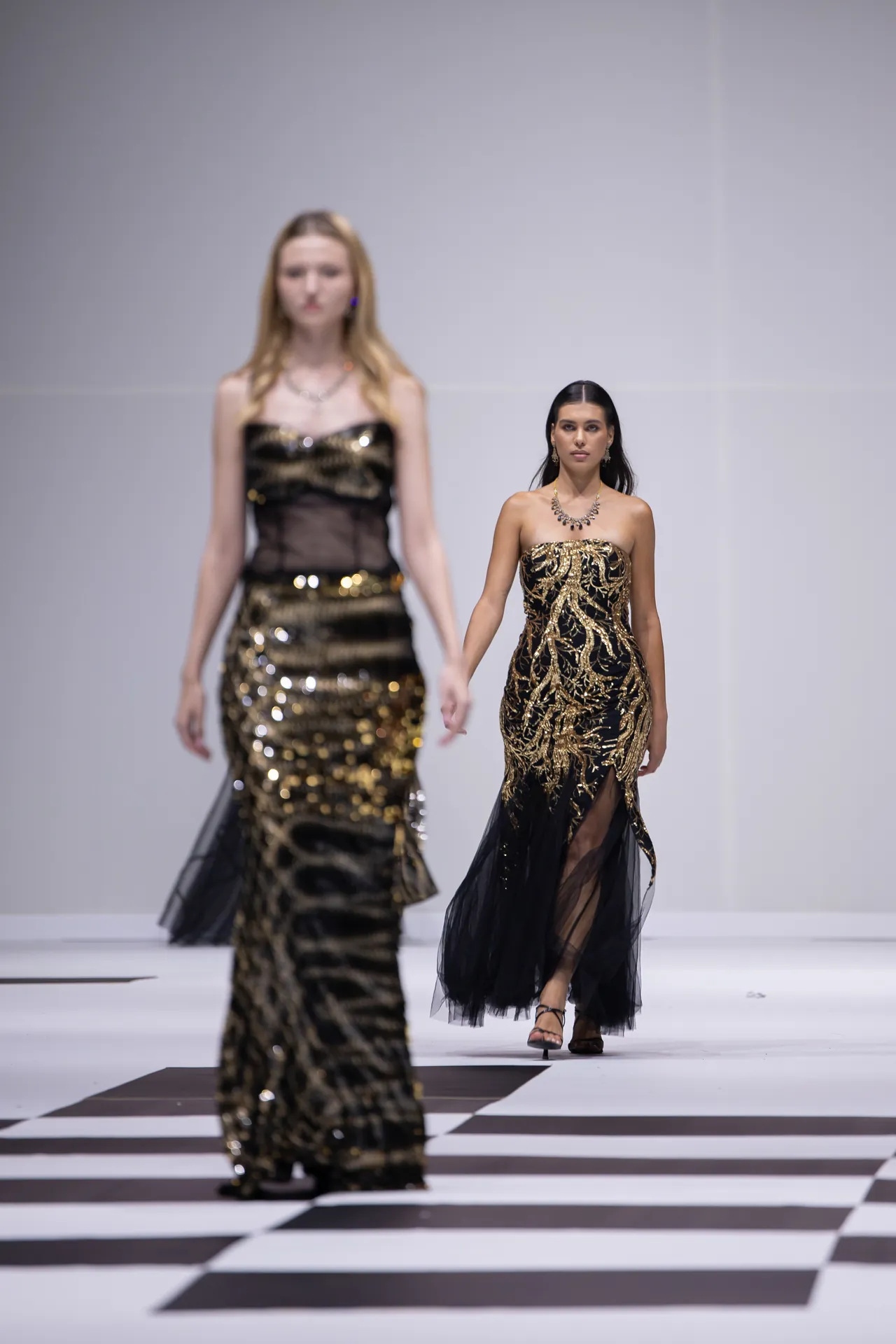 Model wearing a strapless black gown with gold embroidery walks a checkered runway; another similarly styled model appears blurred in the background.