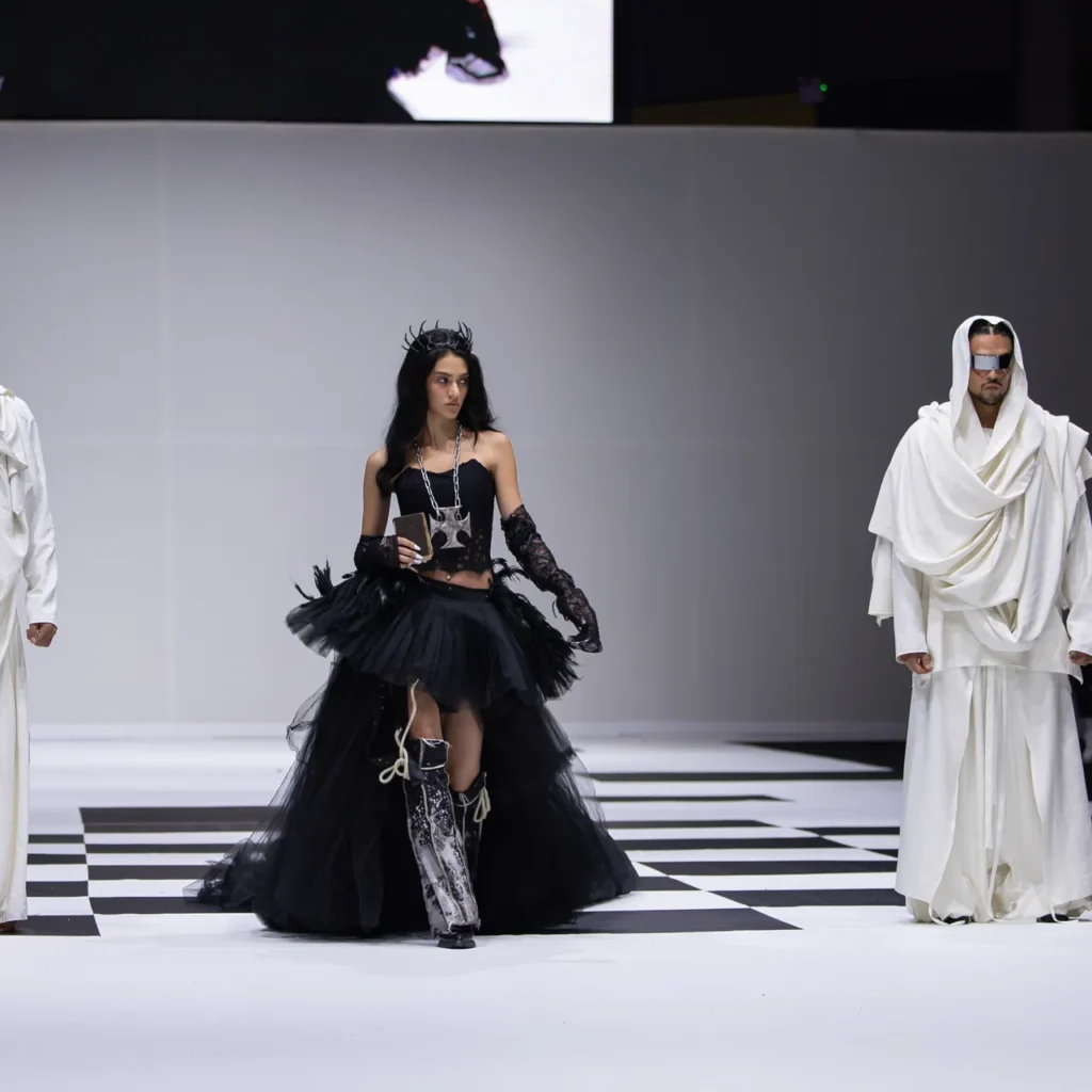 Runway fashion show with three models: center model in a black tulle dress and crown, flanked by two models in white draped outfits and sunglasses on a black-and-white checkered runway.