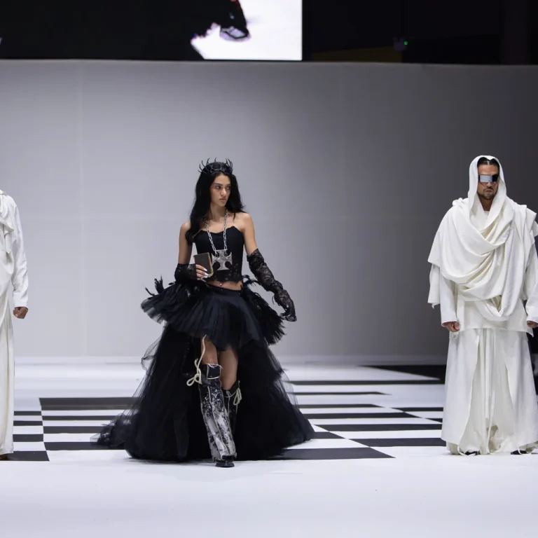 Runway fashion show with three models: center model in a black tulle dress and crown, flanked by two models in white draped outfits and sunglasses on a black-and-white checkered runway.