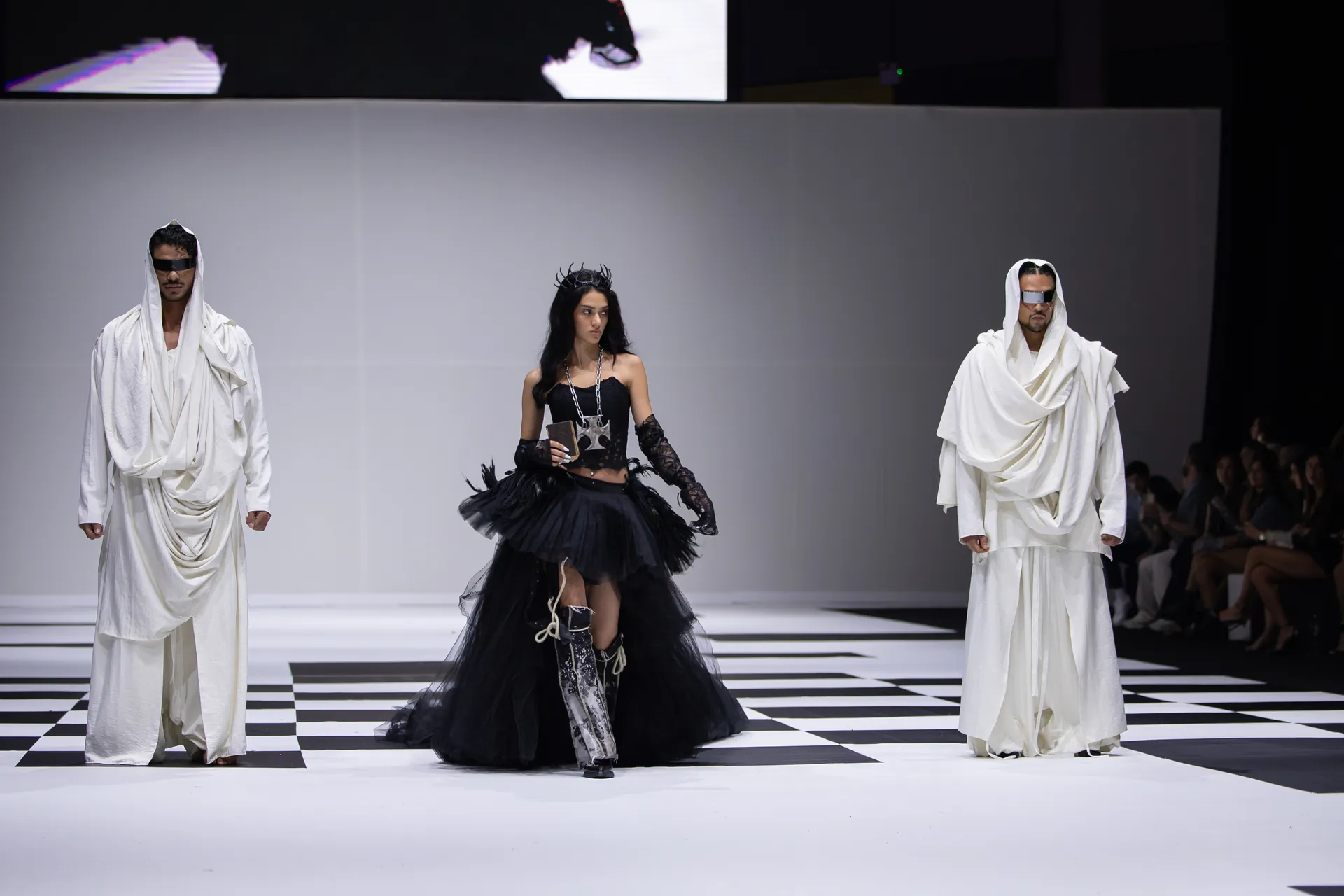 Runway fashion show with three models: center model in a black tulle dress and crown, flanked by two models in white draped outfits and sunglasses on a black-and-white checkered runway.
