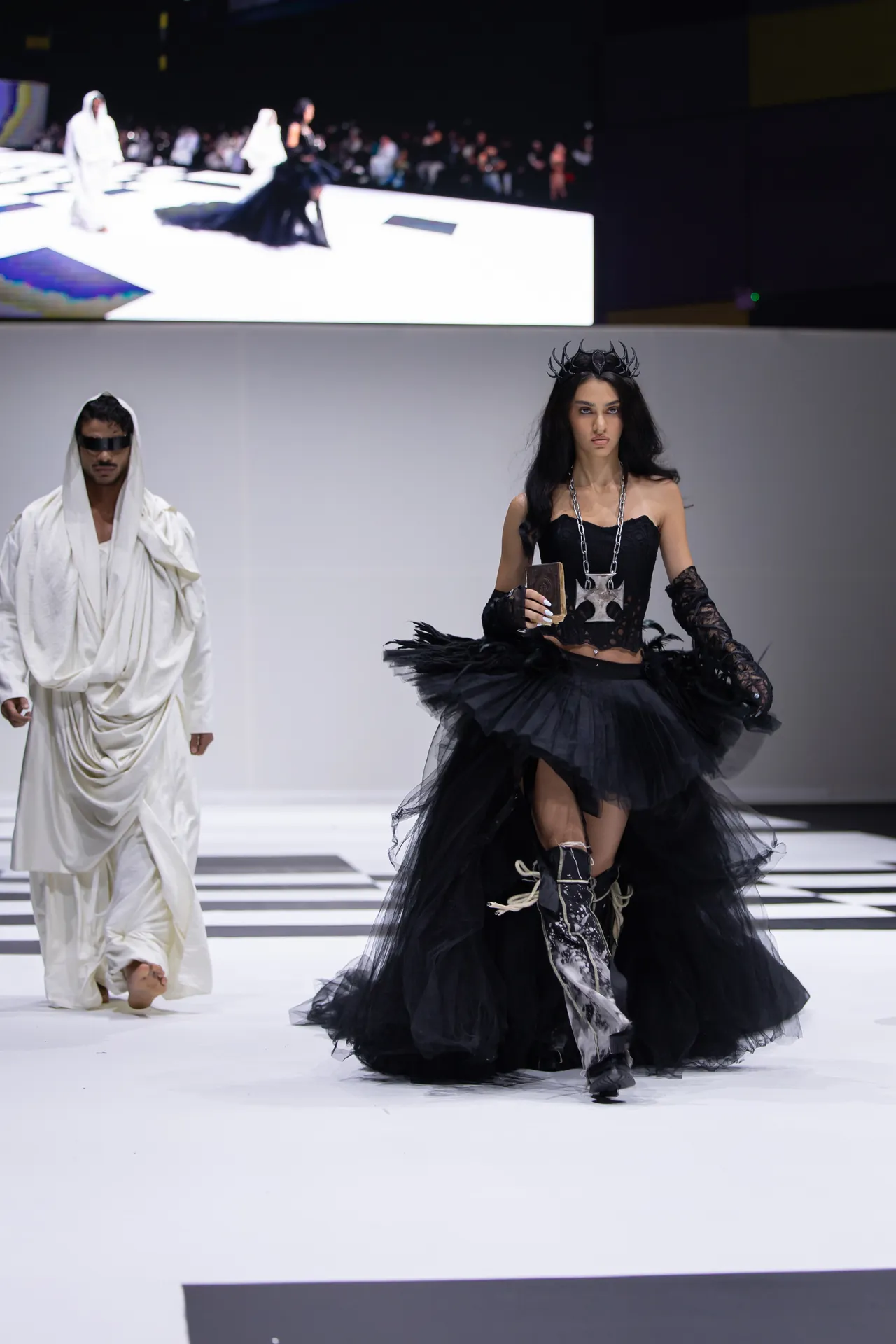 Model in a dramatic black Gothic-inspired gown with a crown walks the runway, holding a small prop, while a second model in a white draped outfit stands nearby on a fashion-show catwalk with a blurred audience in the background.