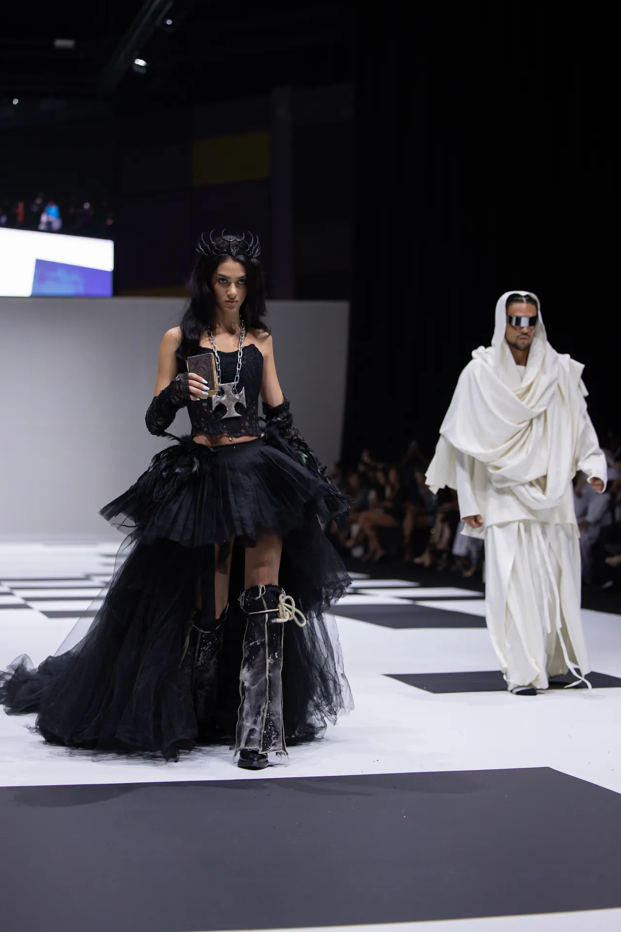 Model on a runway wearing a black gothic gown with a horned headpiece and large tulle skirt; another model in white draped attire in the background.