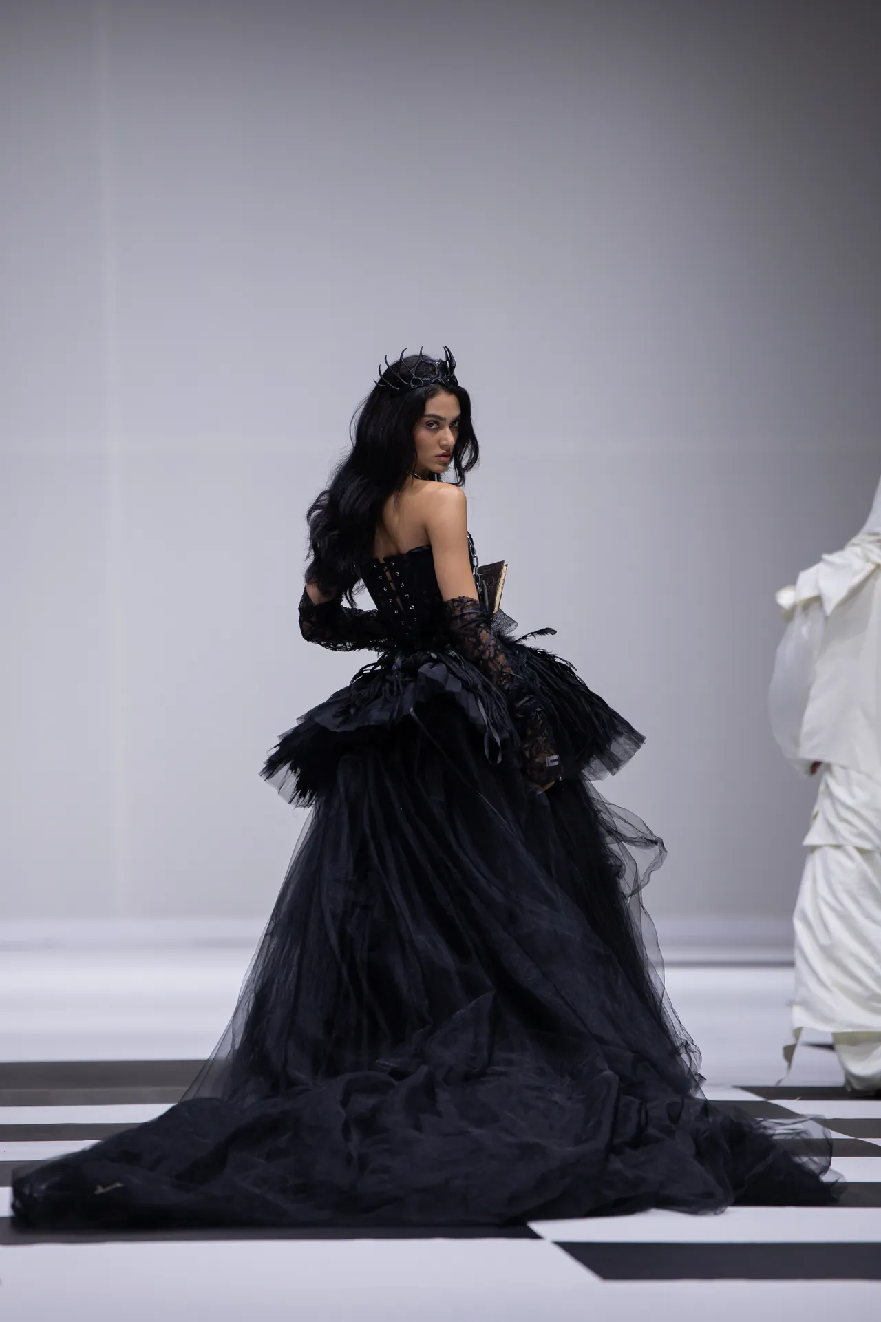Model in a black Victorian-style gown with lace gloves and a dark crown walking the runway, profile turned toward camera.